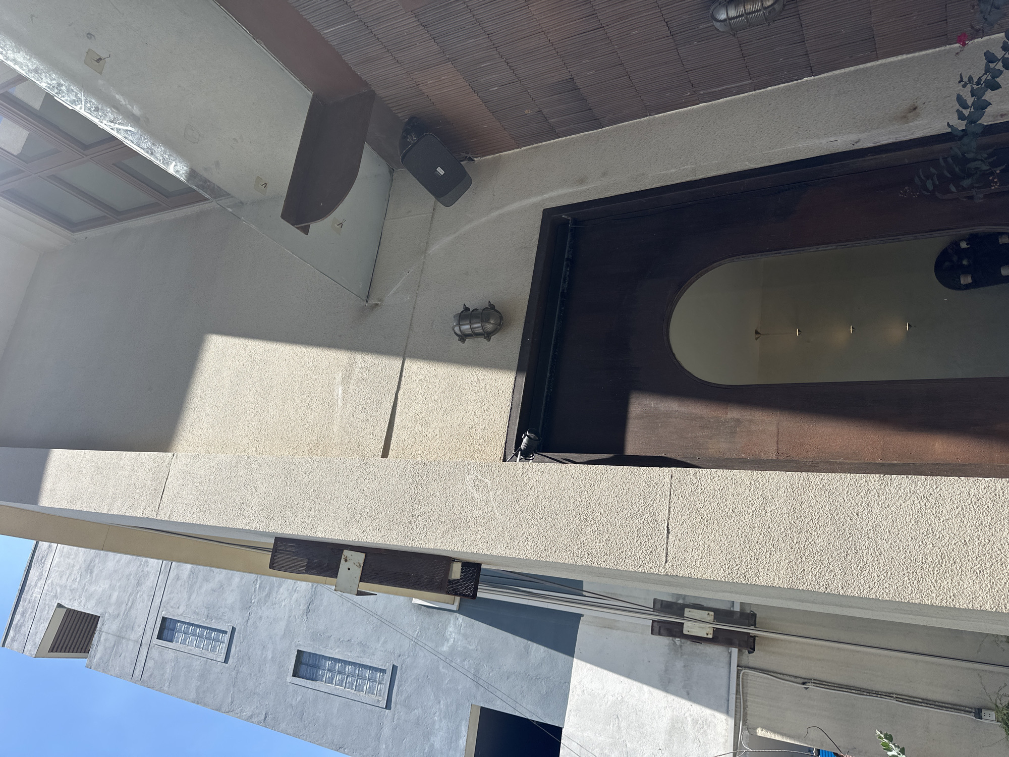 The image shows the exterior of a building with a light beige, textured facade and a dark arched doorway. Above the doorway, a small silver light fixture is mounted to the wall. To the right of the doorway, a dark brown, slatted wall is visible, and mounted on the beige facade above this is a black speaker. The building is adjacent to a taller, light grey building with windows. Sunlight casts shadows across the facade. The scene appears to be in a city environment, possibly Đà Nẵng, Vietnam, given the architectural style and materials.
