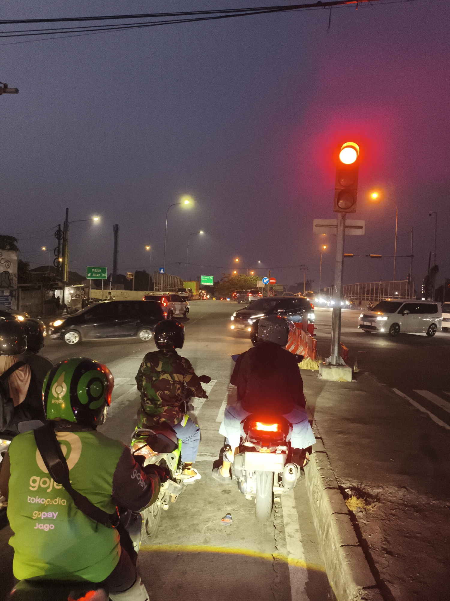 At dusk in Depok, Indonesia, a traffic light turns red, halting several motorcycles and cars on a busy street. The sky is a deep, hazy purple, and the streetlights cast a dim, orange glow.
In the foreground, three motorcyclists, all wearing helmets, are stopped. The rider on the left wears a bright green Gojek jacket, with the Gojek logo prominently displayed. The rider in the middle wears a camouflage jacket, and the rider on the right, facing away from the camera, wears a dark jacket and blue pants. Their motorcycles have illuminated taillights.
Beyond the stopped motorcycles, several cars and other motorcycles are also waiting at the intersection. Two green road signs are visible on poles; one reads "MASUK Jalan Tol," indicating an entrance to a toll road. The road is paved, with a concrete curb and a narrow strip of grass on the right. Overhead, power lines crisscross the sky. The overall atmosphere suggests a typical evening commute in a bustling Indonesian city.
