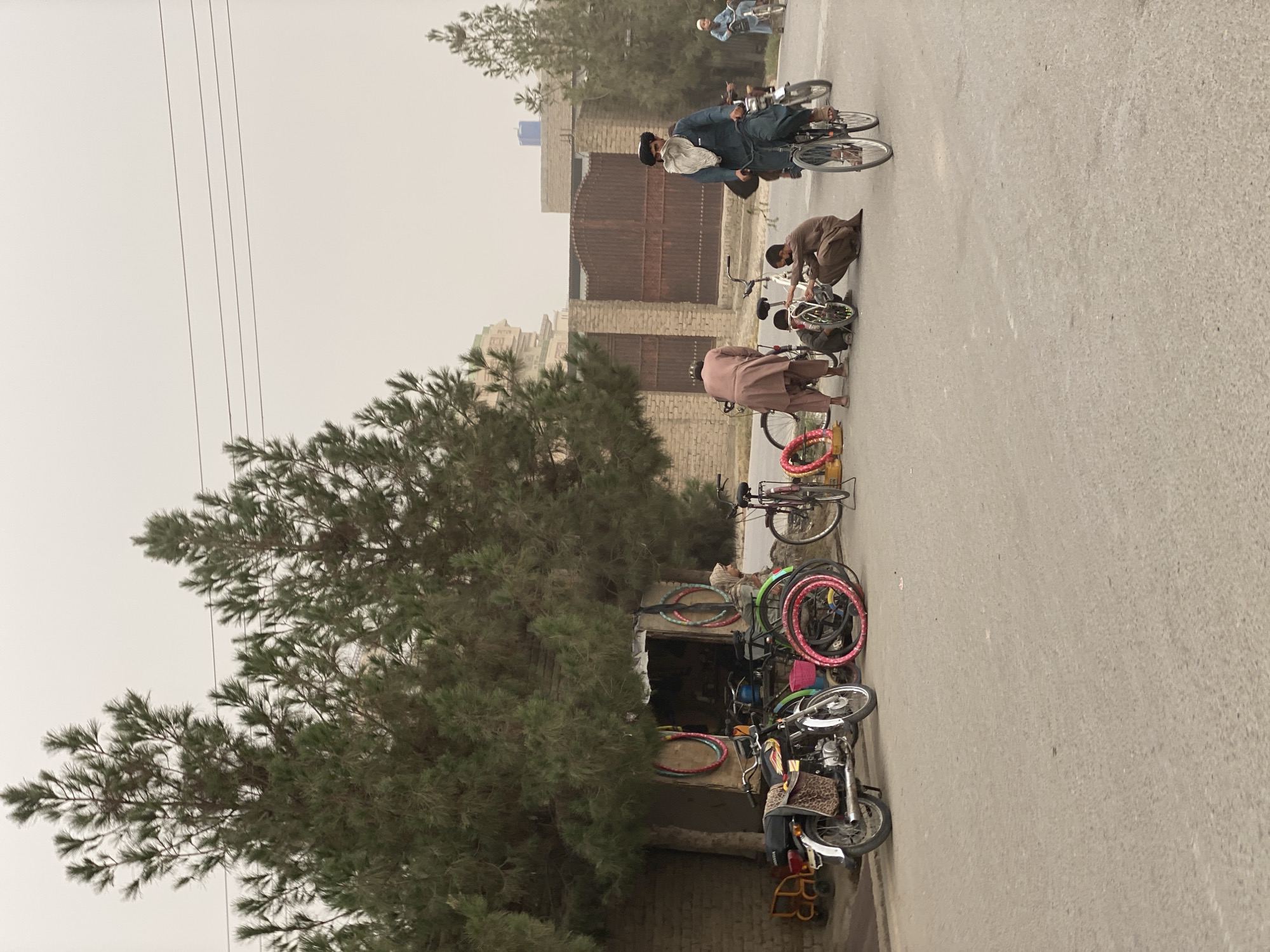 The image depicts an outdoor scene in Kandahar, Afghanistan, with a street lined by buildings and trees. Several people are present, most of whom are interacting with bicycles. In the foreground, multiple bicycles are parked or displayed in front of a small shop or stall. Some of these bicycles have brightly colored rims. Two men in traditional clothing are crouched down, seemingly working on or examining a bicycle. Further down the street, another man is riding a bicycle. In the background, a few more individuals can be seen, one of whom is on a motorcycle.
The setting appears to be a marketplace or a street with local businesses. The architecture suggests a Middle Eastern or Central Asian location, consistent with the provided context of Kandahar. The weather appears to be overcast or hazy, as indicated by the muted sky and diffused light. The overall atmosphere seems to be one of everyday activity, with people engaged in transportation and commerce. No visible text is present in the image. The scene is captured in a vertical orientation, with the ground and street running from the bottom to the top of the frame.