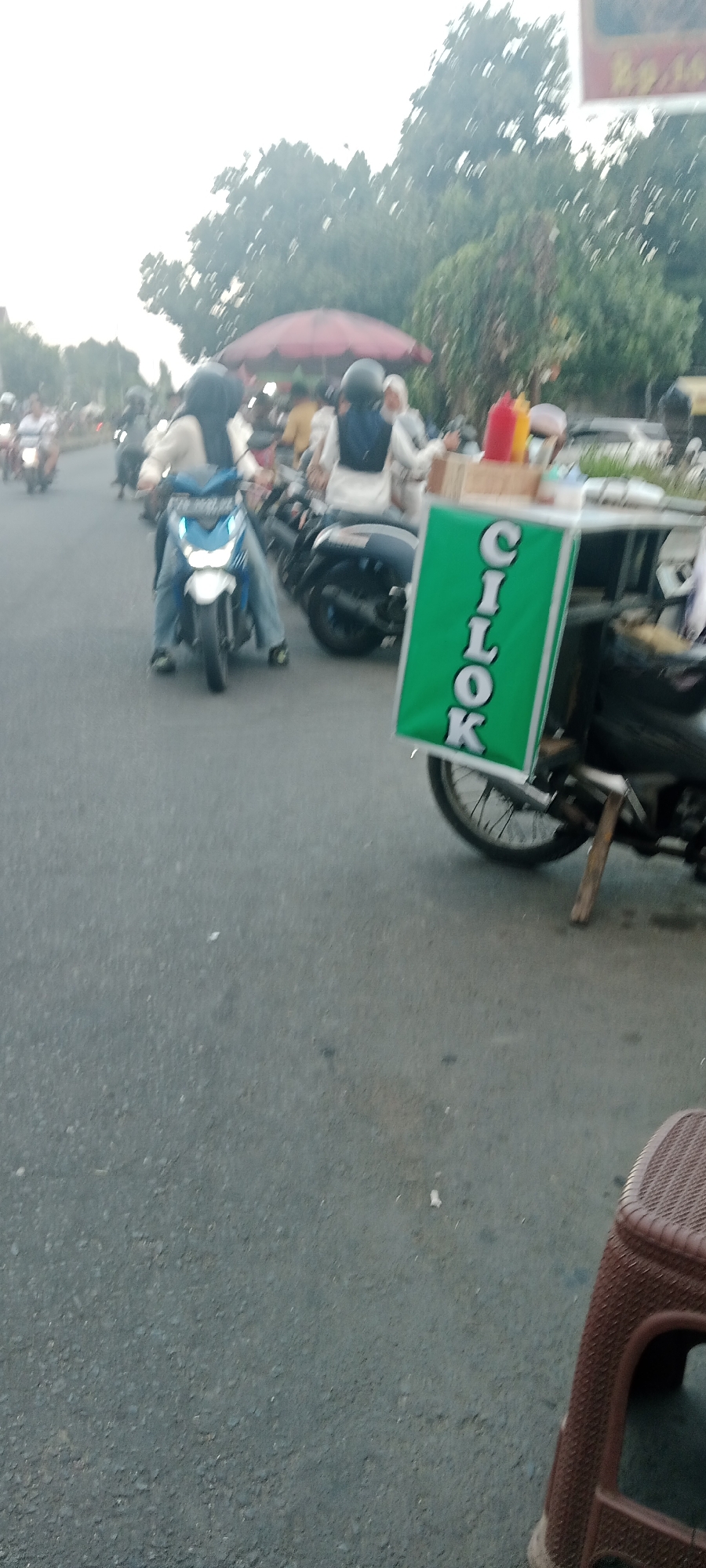 A group of people are riding motorcycles on a paved road in Ngemplak, Indonesia. The image is blurred due to motion. In the foreground, a green sign with white lettering that reads "CILOK" is attached to the side of a motorcycle. There are two bottles of sauce, one red and one yellow, on top of a wooden box on the motorcycle. In the background, trees line the road, and a red umbrella is visible. The sky is overcast.