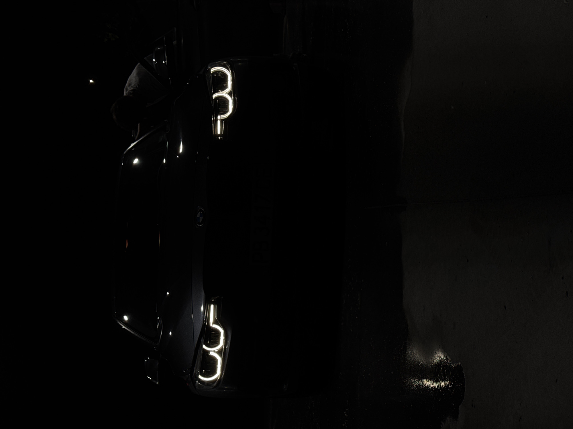 The image shows a dark, low-light scene featuring a car with distinctive illuminated rear lights. The car appears to be a BMW, judging by the logo visible near the top. The lights are turned on, and their reflection is visible on a wet surface. A shadowy figure is seen near the front of the car. The scene appears to be shot in Plovdiv, Bulgaria. Further details about the specific location within Plovdiv, the time of day, and the precise make and model of the car cannot be determined from the image. The exact nature of the figure's interaction with the car is also unclear.