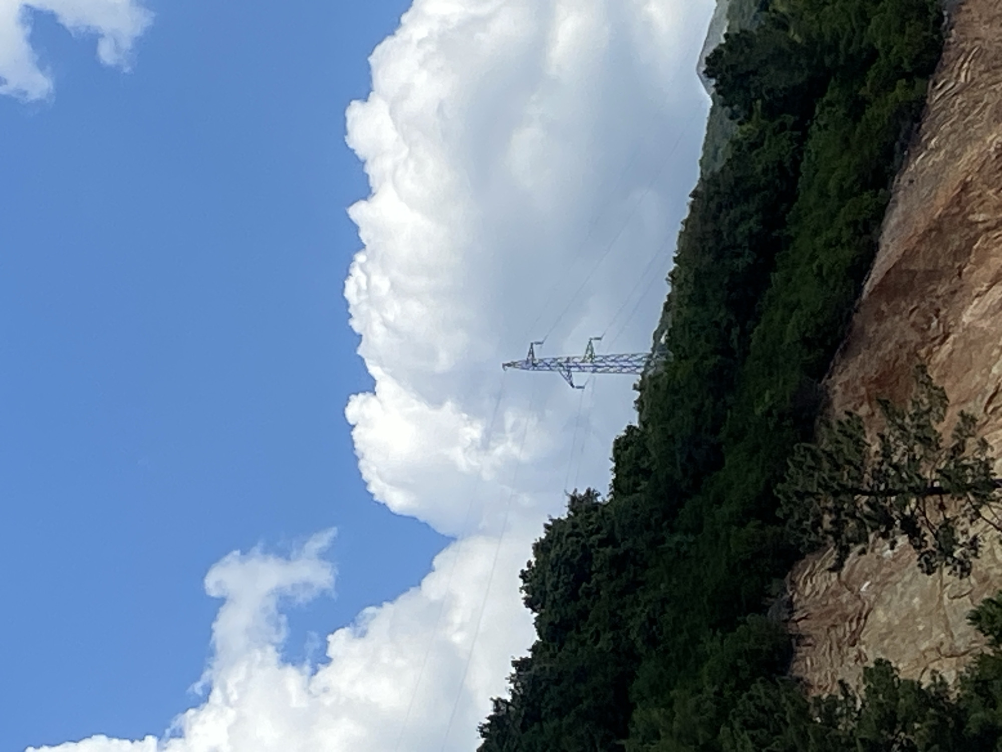 The image captures a scene in Budva, Montenegro. The sky is predominantly blue with scattered white clouds. A large, puffy white cloud dominates the upper-center of the frame. An electrical power transmission tower stands partially obscured by the cloud and a treed mountainside to the right. The mountainside is covered in dense green foliage with exposed patches of light-brown rock.