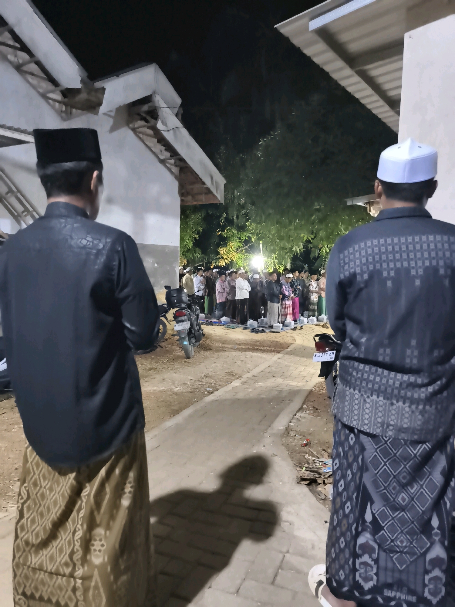 In the city of Pakamban Daya, Indonesia, at night, a group of Muslim men are gathered outdoors, seemingly for a religious observance. Two men are prominently positioned in the foreground, facing away from the camera and towards the group. The man on the left wears a black peci hat and a black patterned shirt with a gold patterned sarong. The man on the right wears a white peci hat and a black patterned shirt with a black patterned sarong that has "SAPPHIRE" written on it.
The group in the background consists of approximately 20-30 men, also wearing peci hats and sarongs. They are standing in rows, facing forward, with several prayer mats laid out on the ground. A motorcycle is parked among them. The environment appears to be a courtyard or open area next to a building with a corrugated roof. The lighting suggests it is nighttime, with some artificial lights illuminating the scene. The ground is made of dirt and paving stones. The overall atmosphere is one of communal gathering and prayer.