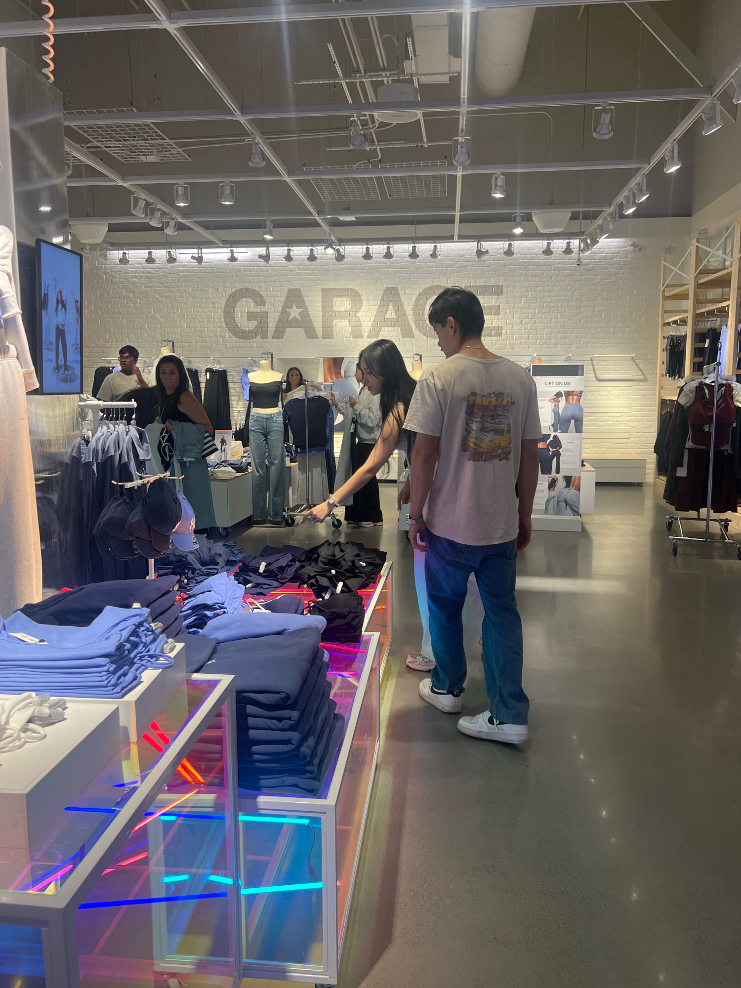The image captures an interior view of a clothing store, identified by the prominent "GARAGE" logo on a white brick wall. The store appears to be located in Vaughan, Canada. In the foreground, neatly folded stacks of blue and navy clothing are displayed on a multi-level retail fixture with colorful, illuminated shelving. To the right of this display, a young woman in a black top and jeans is holding a pair of jeans, looking at them while a young man in a light-colored t-shirt and blue jeans stands beside her, looking in the same direction. Behind them, mannequins are dressed in contemporary fashion, and racks of clothing are visible. Further in the background, other shoppers and store employees are present, interacting with merchandise. The store's ceiling features a grid system with lighting fixtures, and the floor is a polished concrete.