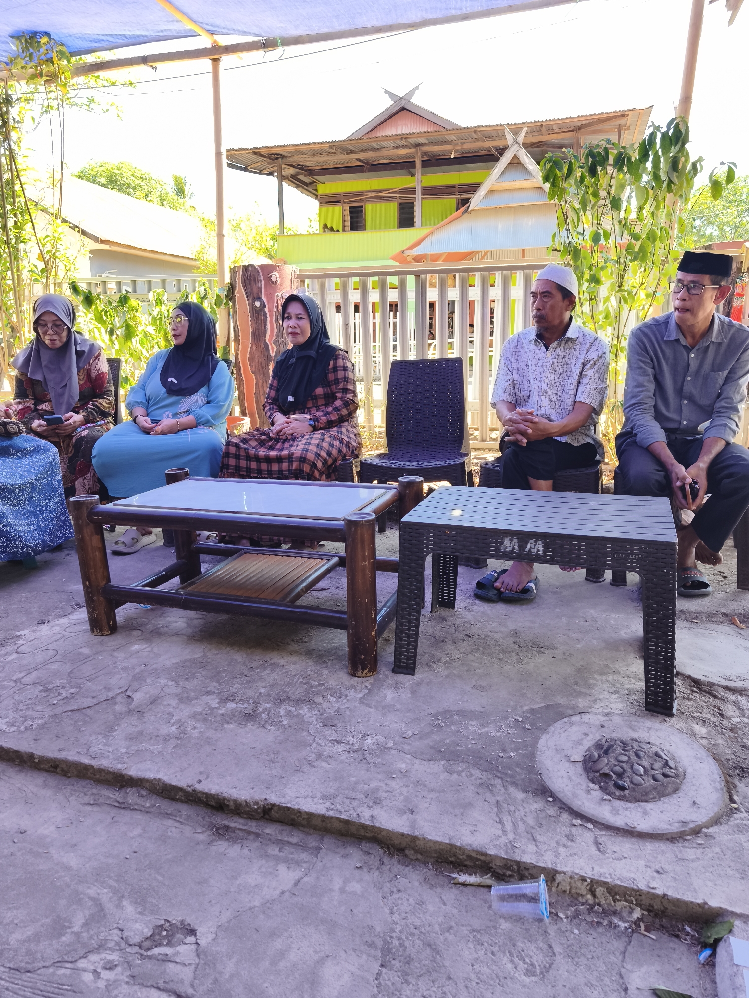 In Tonasa, Indonesia, a group of four adults, two women and two men, are seated in a line outdoors. They are positioned in front of a building with a green and yellow facade and a corrugated metal roof. The women, on the left, are wearing hijabs and traditional clothing. The man in the middle is wearing a white skullcap and a patterned shirt, while the man on the right is wearing glasses and a grey shirt. Two low tables, one made of dark wood with a glass top and the other a dark plastic coffee table with the inscription "MM," are placed in front of them. The ground is paved with concrete, showing signs of cracking and wear. Vegetation, including bamboo-like stalks and leafy plants, grows around the seating area. A blue plastic cup lies discarded on the ground near a circular concrete fixture with stones. The scene suggests a casual gathering or meeting.