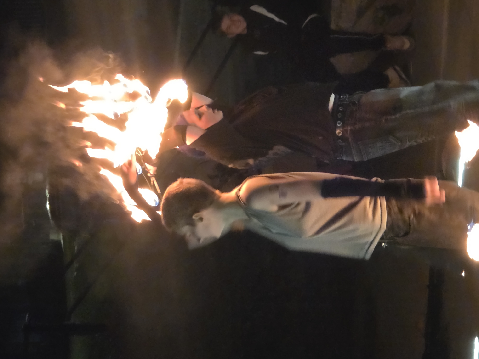 A group of people are performing with fire at night. Two individuals are in the foreground, with one on the left facing away from the camera and holding a flaming object above their head. This person is wearing a sleeveless shirt and jeans. The person on the right is facing forward and also holding a flaming object. This individual is wearing a black top and jeans. In the background, another person is visible, appearing to be watching the performance. The scene is dimly lit by the fire and some artificial lights. The location is not specified.