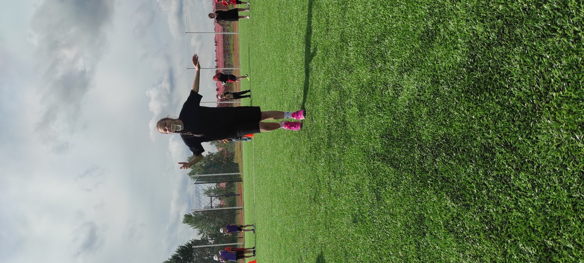 In Noviy, Russia, a young girl with braided hair is standing on a green turf field, possibly a sports ground, with a plastic cup balanced on her teeth. She is wearing a black t-shirt, black shorts, and bright pink athletic shoes. Her arms are outstretched, and she appears to be participating in some sort of outdoor activity or game. The background shows a chain-link fence, trees, and buildings, suggesting an urban or suburban setting. There are other people, including children, in the background, some of whom are also engaged in activities. The sky is cloudy, indicating overcast weather.