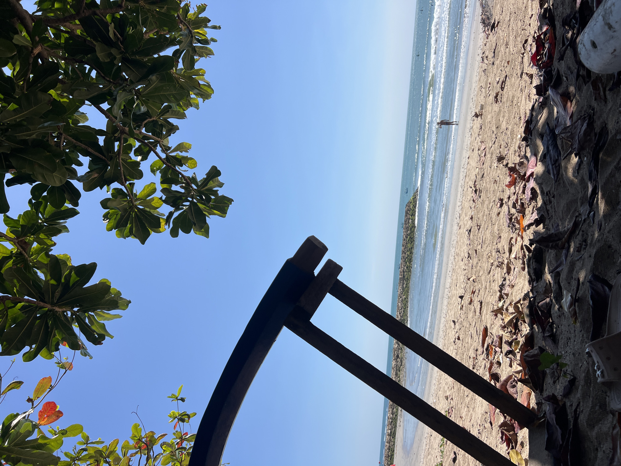 An outdoor, eye-level, vertical shot captures a serene beach scene. In the foreground, dry brown leaves litter the sandy ground, partially obscuring a light-colored sandal. Two dark wooden poles, part of a beach chair or structure, extend diagonally across the frame from the lower left towards the upper right. Above, lush green leaves from a tree hang down, with a few splashes of red and orange among them, contrasting with the clear, bright blue sky.
The midground reveals a sandy beach with gentle waves lapping the shore. A breakwater, constructed of grey rocks, runs parallel to the coastline. In the distance, a lone person stands near the water's edge, appearing to be looking out at the ocean. The sea stretches out to the horizon, dotted with a few small boats. The overall impression is one of tranquility and natural beauty. The scene is set on the beach in Kuta, Indonesia.