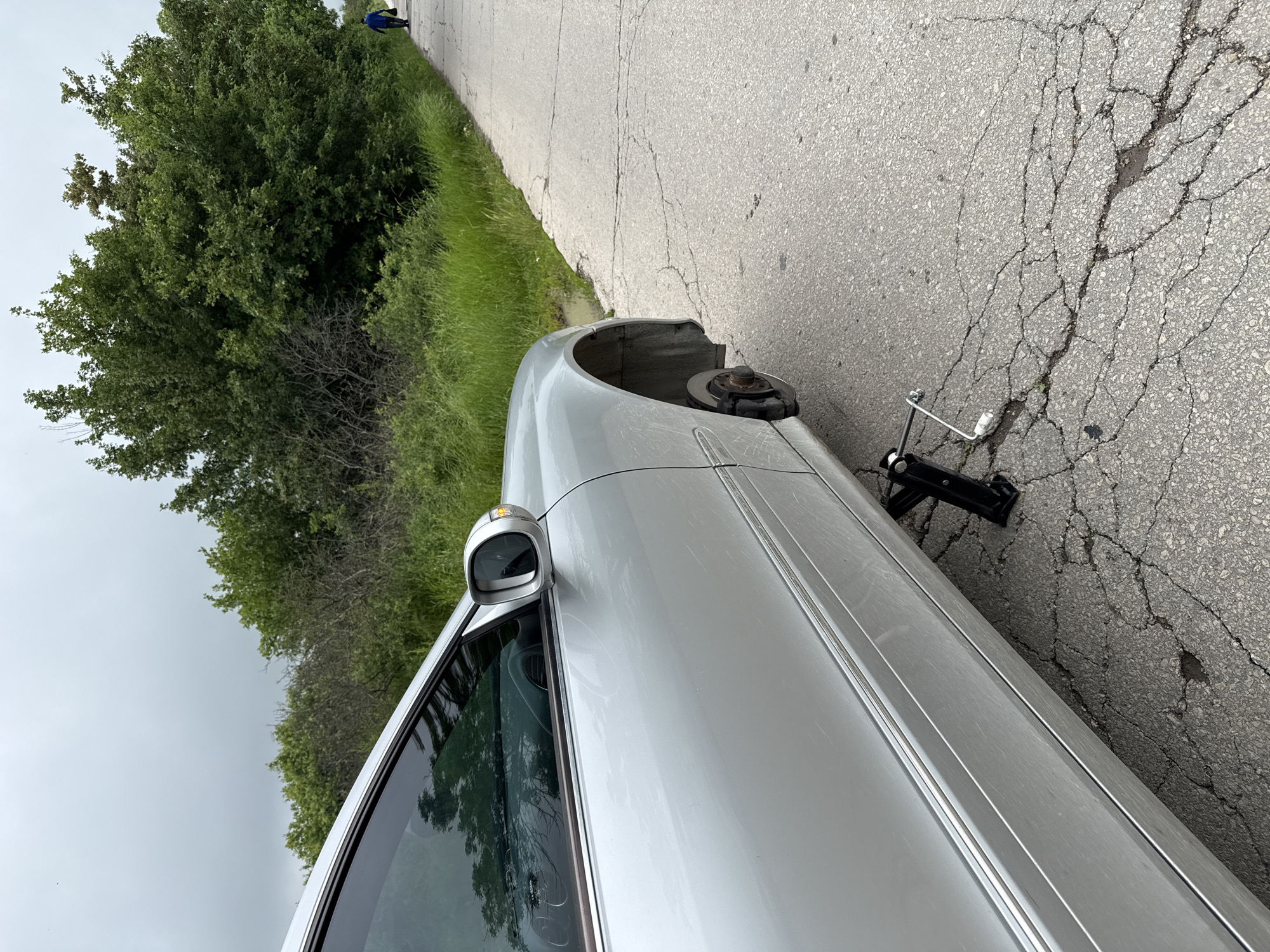 The image displays a silver sedan, possibly a Mercedes-Benz, parked on the side of a worn asphalt road. The vehicle has its front passenger-side wheel removed, exposing the brake rotor and caliper. A black car jack with a silver handle is positioned on the cracked asphalt next to the car, indicating a tire change or repair is in progress.
The road surface is gray, showing extensive cracking. Alongside the road is a green grassy verge, transitioning into dense foliage consisting of trees and bushes. The sky is overcast and uniformly light gray. In the far distance, a person dressed in blue is visible on the road, partially obscured by the trees.
The scene suggests a daytime event in a rural or semi-rural setting, likely involving an automotive issue like a flat tire. No visible text is identifiable on the car or other objects. The overall impression is one of a minor roadside inconvenience.