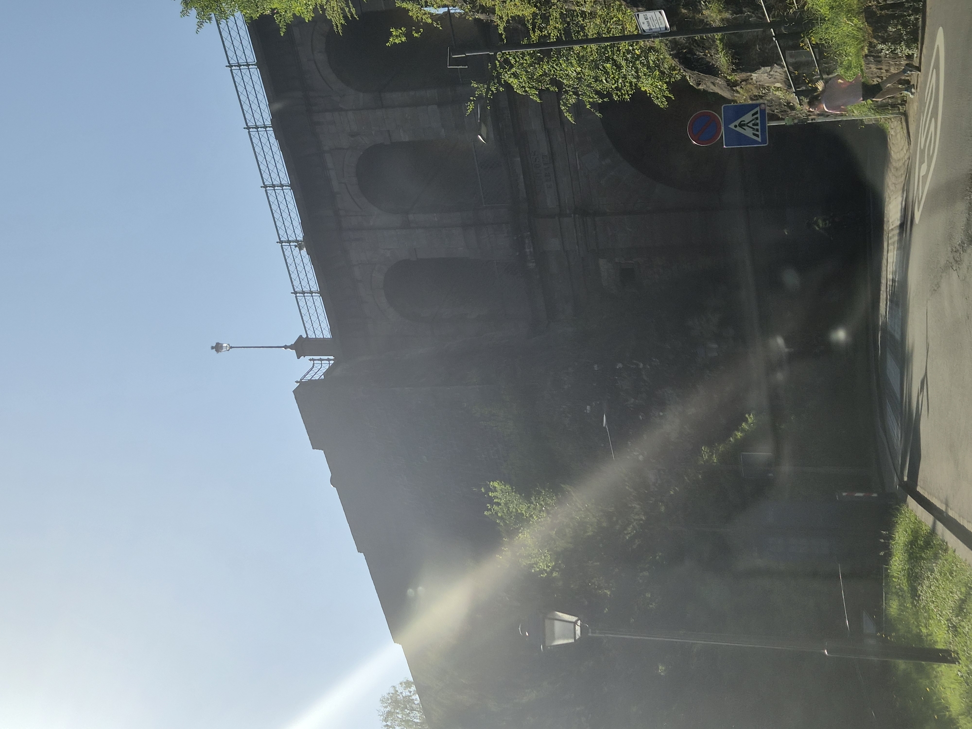 The image depicts a street scene in Luxembourg City, Luxembourg, focusing on a large stone viaduct. The viaduct has multiple arched openings and a metal railing along its top. A streetlamp is visible on the left side of the frame, and sunlight streams in from the upper left, creating lens flare. On the right side of the road, there are two traffic signs: a circular red and blue sign and a blue square sign with a pedestrian crossing symbol. A person is running on the right side of the road near the pedestrian crossing. The road appears to be asphalt and has white markings. Green grass and foliage are present along the sides of the road and on the viaduct structure. The scene suggests an outdoor environment, possibly a park or a well-trafficked area within the city.