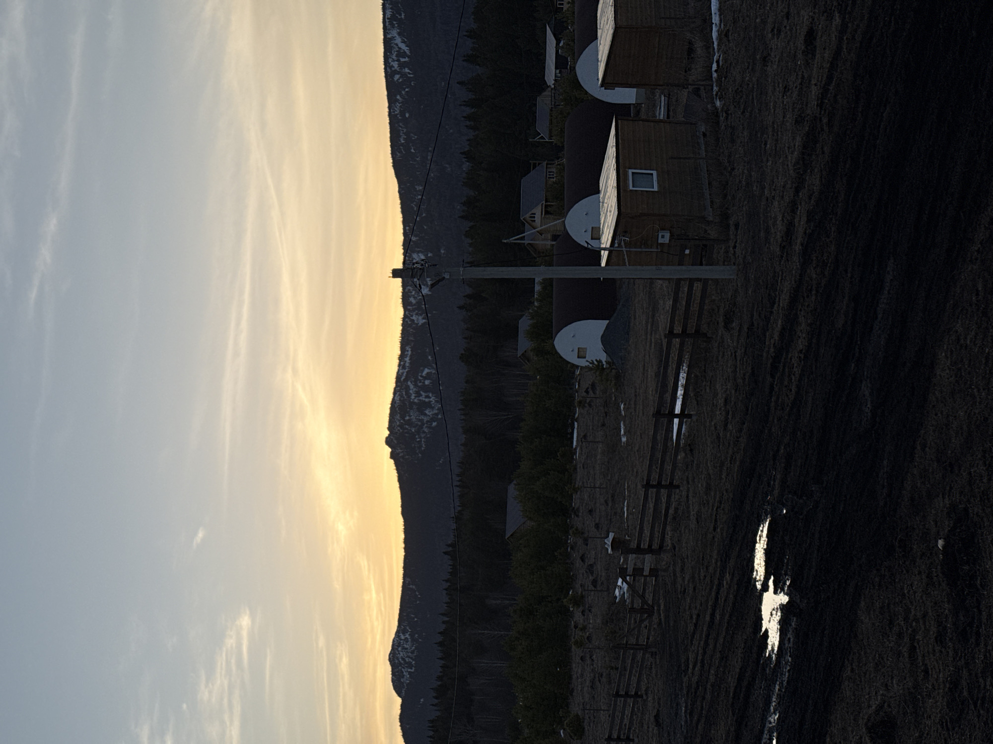 The image depicts a rural or semi-rural scene in Tyulyuk, Russia, at either sunrise or sunset. The sky displays a gradient from pale blue at the zenith to a strong orange-yellow glow at the horizon, with sparse, wispy clouds. In the background, dark, snow-capped mountains form a silhouette against the bright sky, with a dense, dark evergreen forest directly in front of them.
In the midground, a cluster of structures is visible. Several dark, semi-cylindrical buildings, resembling quonset huts with white end caps, are present. A pile of light-colored gravel is situated next to one such structure on the left. Closer to the foreground are two rectangular wooden sheds with light-colored, sloped roofs. The shed on the left features a small, square white-framed window. Further back, nestled among trees, are several traditional wooden houses with pitched roofs. An electrical utility pole stands vertically in the center-right, with power lines extending horizontally across the scene. A wooden fence line runs across the midground from left to right, featuring a gate section.
The foreground consists of dark, uneven ground, characterized by dry, sparse vegetation and significant areas of exposed dark mud. A large, irregular puddle in the left-center foreground strongly reflects the bright orange-yellow sky. Patches of residual snow are visible on the ground near the fence line and scattered among the distant structures. No individuals or specific actions are depicted.
