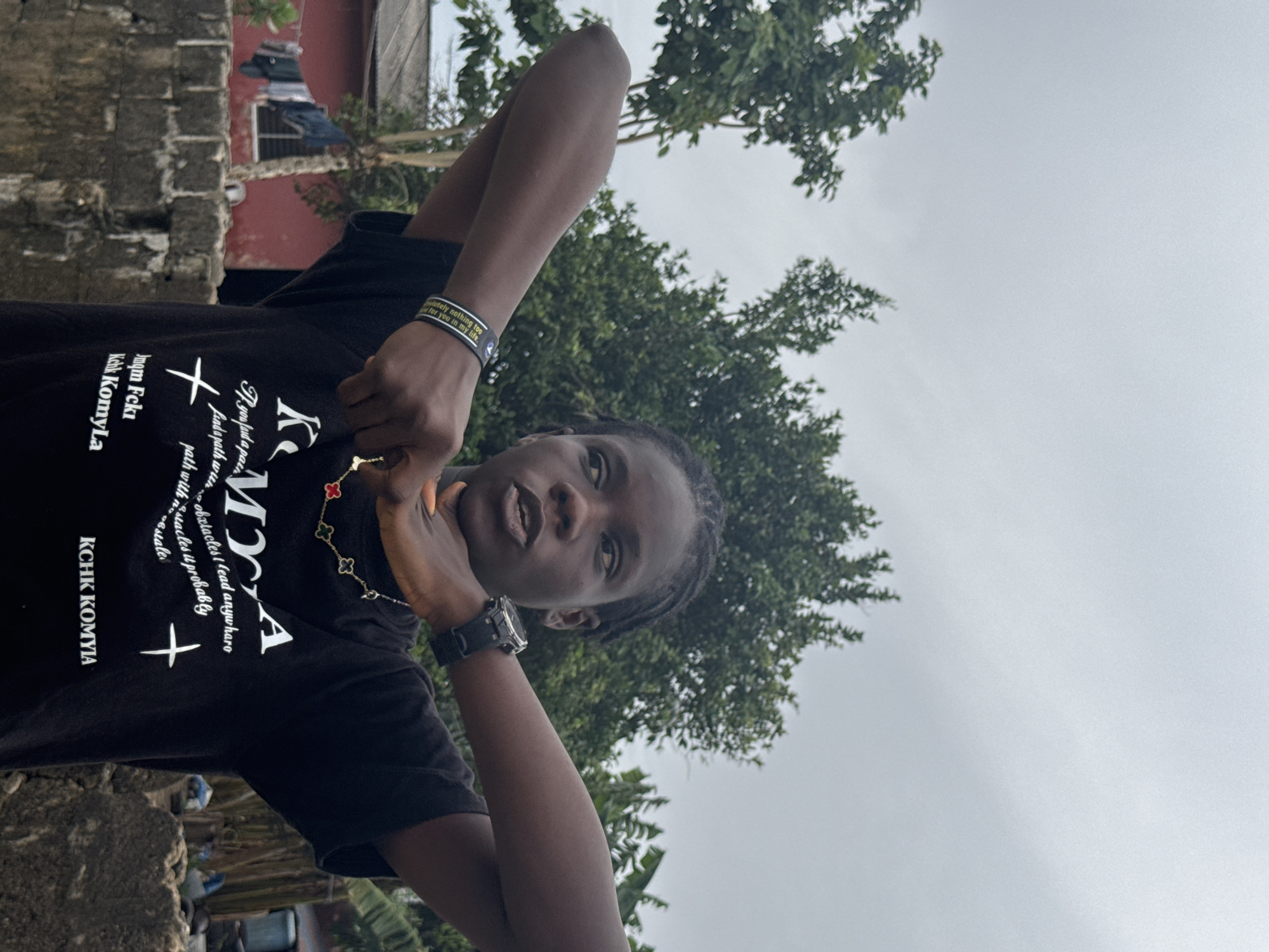 A photograph captures a person in an outdoor setting, with a partial view of a building and greenery. The person is wearing a dark-colored t-shirt with writing, a bracelet, a necklace with a floral design, and a watch. They are holding their hands near their neck. The sky is overcast. Part of a brick wall is visible. There is evidence of urban development, suggested by the building and possibly a car. The exact location is uncertain.