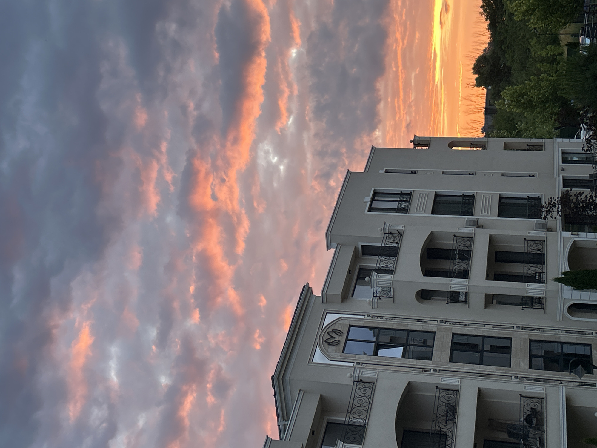 The image shows a modern apartment building in Plovdiv, Bulgaria, at sunset. The sky is filled with dramatic, colorful clouds, ranging from deep grays to vibrant oranges and pinks. The building itself is light-colored with multiple balconies, ornate railings, and large windows. Green trees are visible in the background, some of which are silhouetted against the bright sky. The overall impression is one of an urban landscape bathed in the warm, dramatic light of a setting sun.