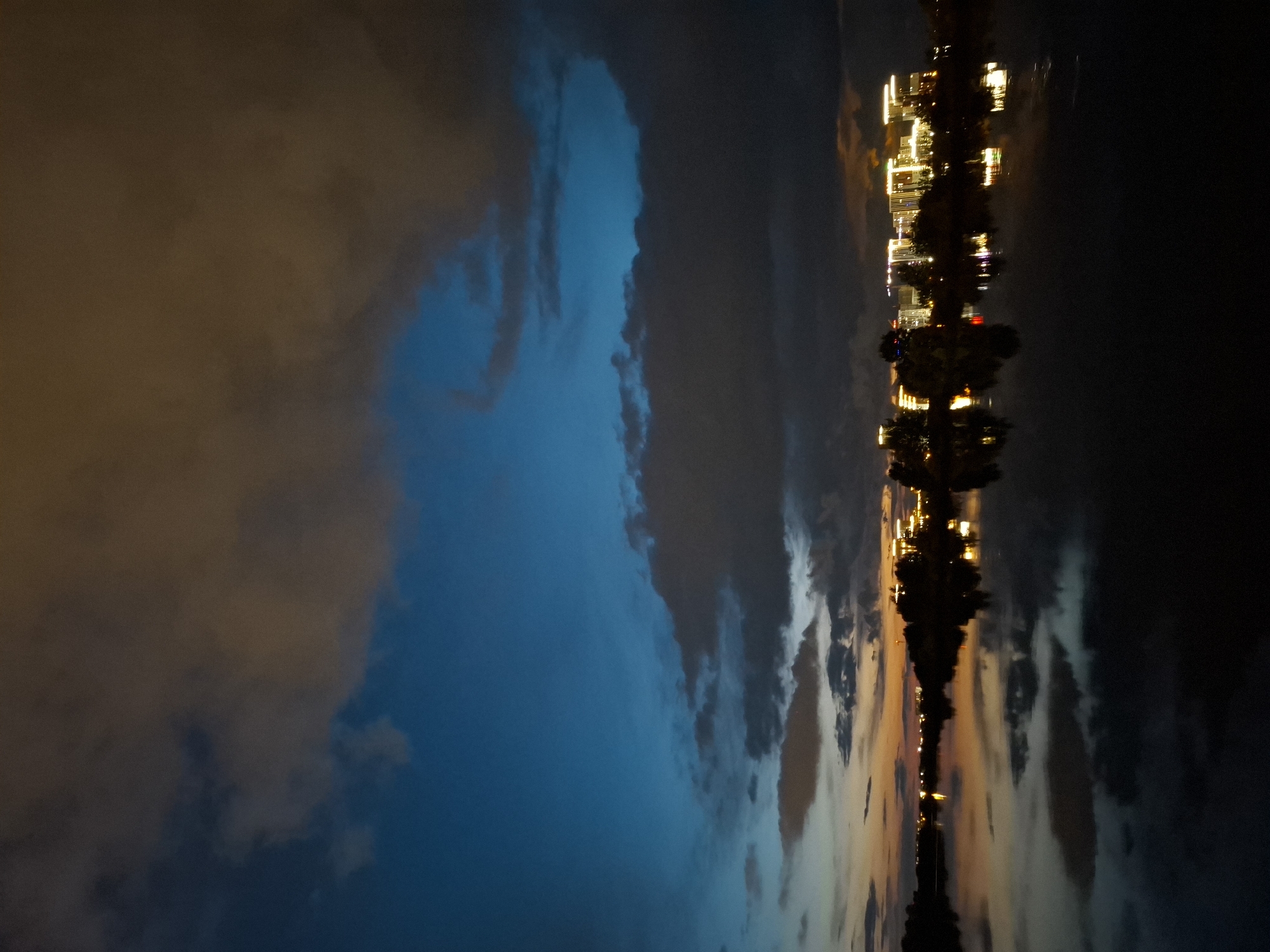 The image depicts a twilight scene over a body of water, likely a river or lake, with a cityscape in the background. The sky is a gradient of deep blue and dark gray clouds, with pockets of orange and yellow suggesting the last vestiges of sunset or artificial lighting. The water in the foreground reflects the sky and the illuminated buildings, creating a symmetrical composition.
The cityscape features a cluster of modern buildings, illuminated by numerous lights, casting a warm glow. These structures appear to be residential or office buildings, characterized by their rectangular shapes and illuminated windows. Trees are interspersed among the buildings, silhouetted against the lights.
The body of water appears calm, with smooth reflections, indicating a lack of significant wind or wave activity. The overall atmosphere is serene and urban, capturing the transition from day to night in a developed area.
Given the context of Tyumen, Russia, this scene could be representative of the waterfront areas, particularly those with newer developments. For instance, the embankment of the Tura River in Tyumen, especially the sections with modern apartment complexes and business centers, often presents such a view at dusk. Areas like the residential complexes near the bridge of Lovers or the city center's riverside developments could offer similar panoramic vistas. The presence of multiple lit buildings arranged along the horizon strongly suggests a developed urban waterfront.