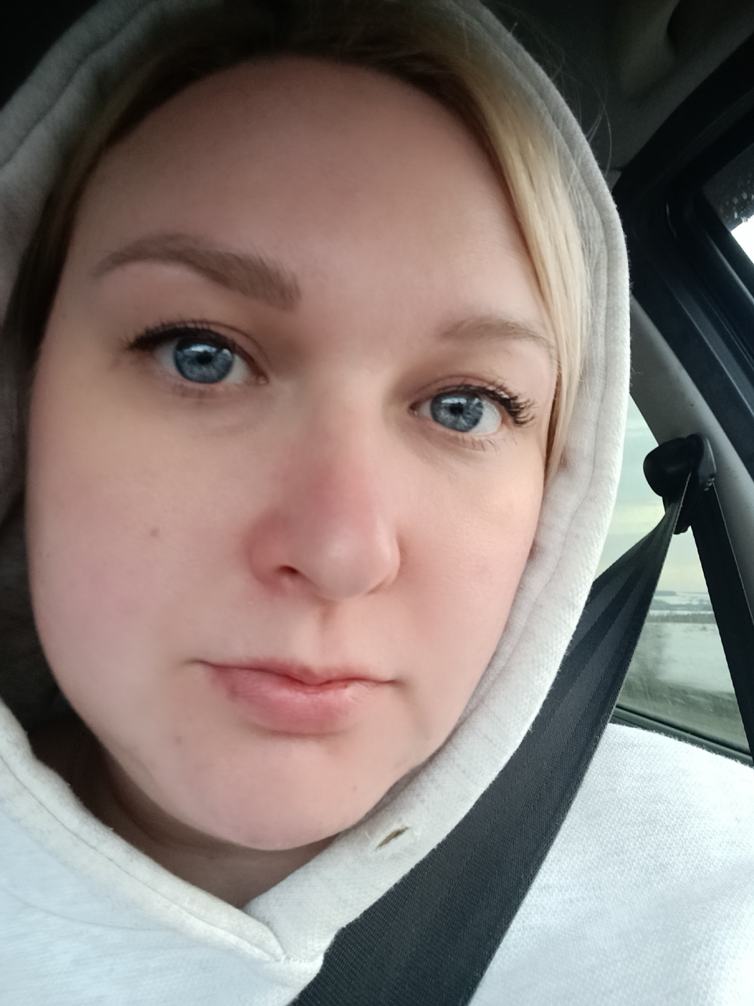 A close-up selfie depicts a Caucasian woman with light blonde hair, framed by a white hooded sweatshirt and a visible black seatbelt in a vehicle. Her eyes are a pale blue, and she has a mole on her left cheek. Through the car window, a snowy landscape is visible, suggesting an outdoor environment. The location is indicated as Koliushevo, Russia.