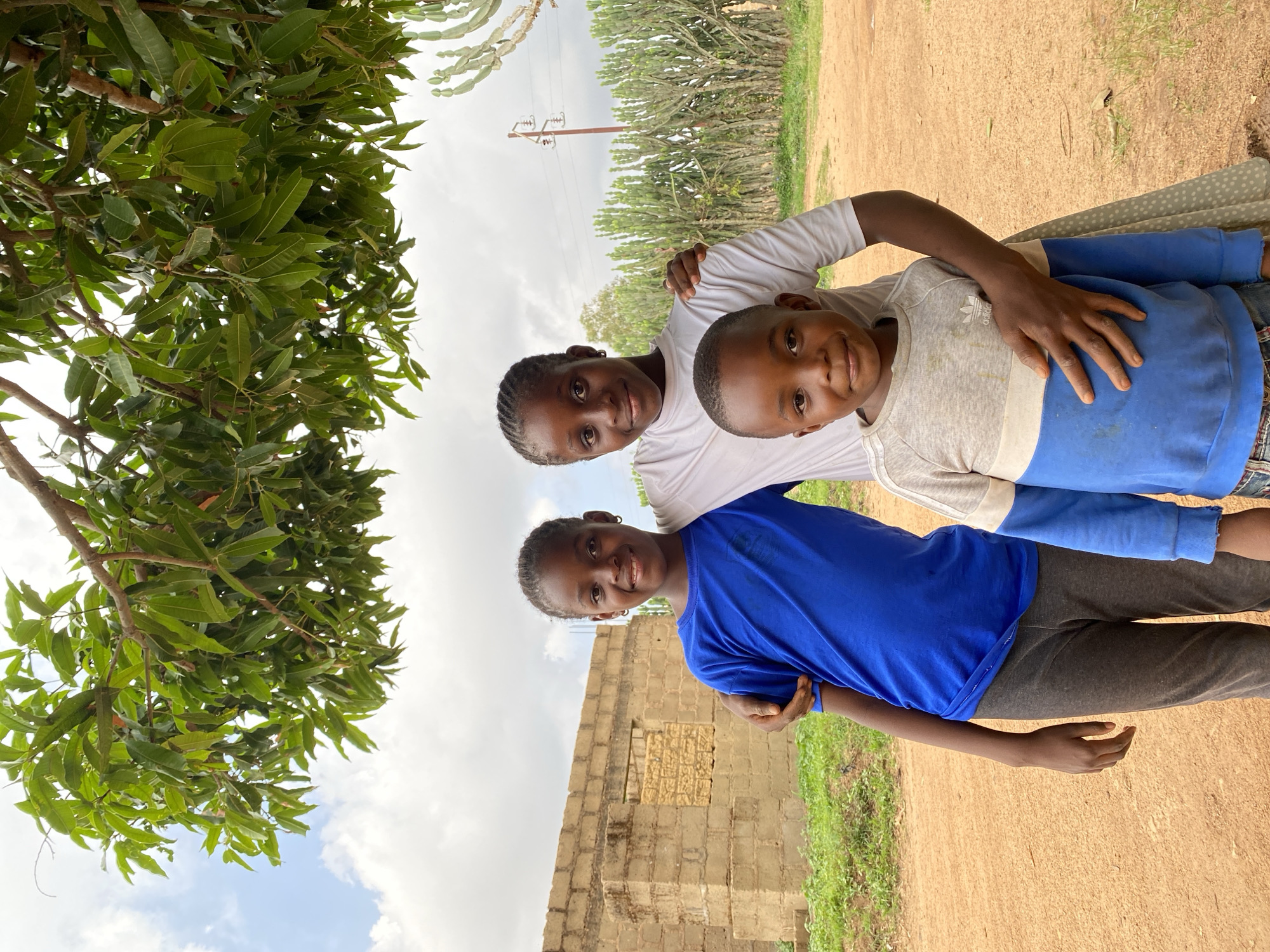 Three children are posing for a photograph outdoors in Vom, Nigeria. A girl wearing a white t-shirt stands at the top, her arm around a younger child wearing a grey and blue sweatshirt. Below them, another child wearing a blue shirt and grey pants is embraced by the girl. The scene is set on a dirt path with green vegetation nearby. Behind the children, there is a partially constructed brick building. A tree with dense foliage is visible on the left side of the frame, and utility poles can be seen in the background. The sky is overcast.