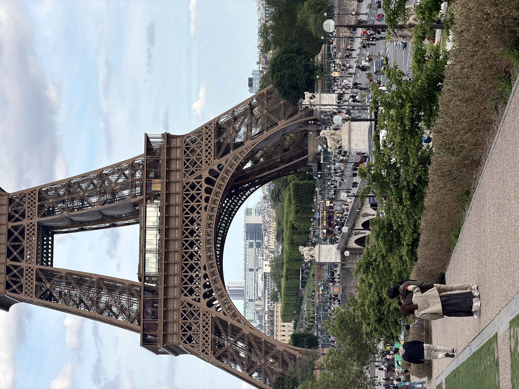 This vertical, eye-level shot shows a portion of the Eiffel Tower in Paris, France. The tower's intricate latticework of brown metal dominates the left side of the image, reaching towards a cloudy sky. In the middle, the structure of the tower arches, revealing buildings and other parts of the city in the background.
On the right, a pathway with people is visible. A couple wearing neutral-colored jackets and dark pants stands near a hedge. Other pedestrians are walking along the path.
Below, a street lined with trees, statues, and lampposts is bustling with activity. There are many people walking along the street and on the bridge. Buildings can be seen in the distance between the trees.