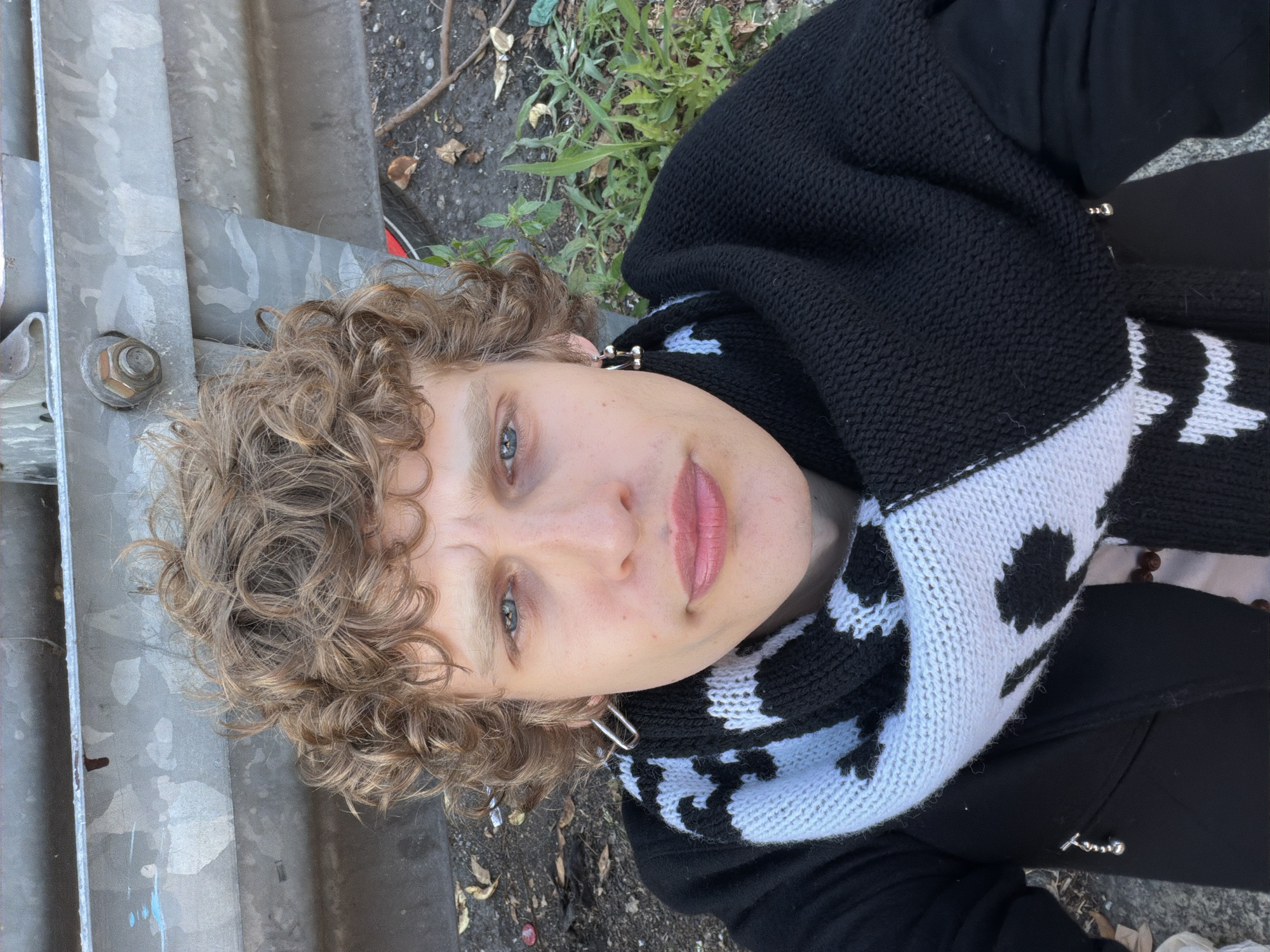 A person with curly blonde hair, blue eyes, and a light complexion is facing the camera, wearing a black and white knitted scarf. The person is outside; a grey metal structure is visible to the left and some green vegetation to the upper right. The person's location cannot be confirmed to be Milan, Italy, based on the visual information provided.