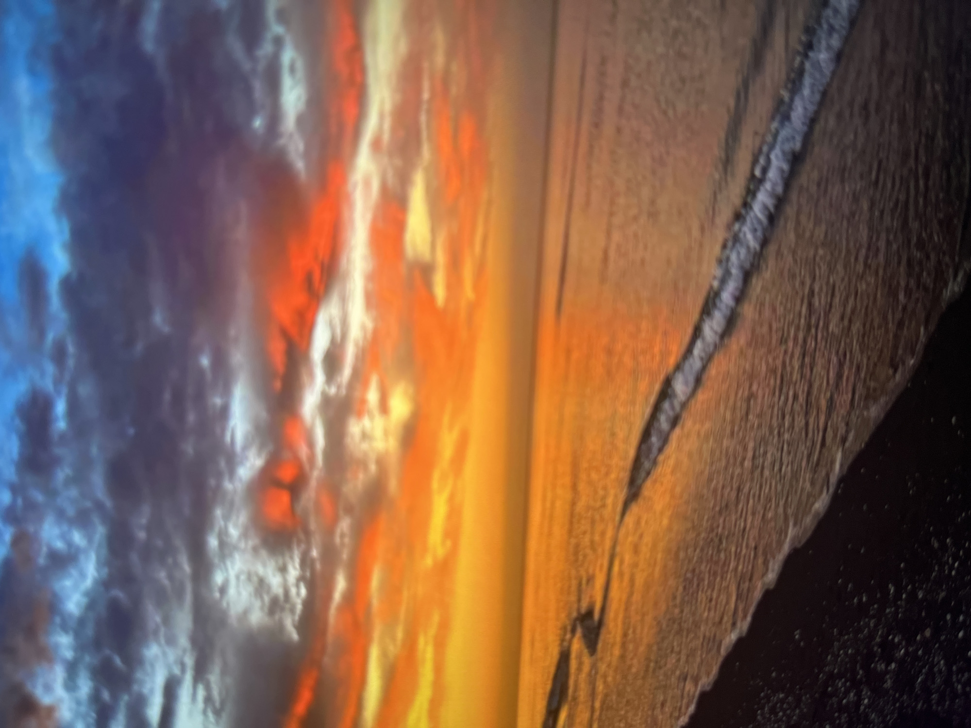 The image captures a vibrant sunset over the ocean, casting a warm, golden glow across the water and the sky. The clouds are painted with dramatic hues of orange, red, and purple, contrasting with the deep blue of the upper sky. A gentle wave breaks on the shore, its white foam tracing a line along the dark, pebble-strewn beach. The scene is serene and evocative of the natural beauty found in coastal regions. The location is the beach in Cirahayu, Indonesia, known for its stunning sunsets.