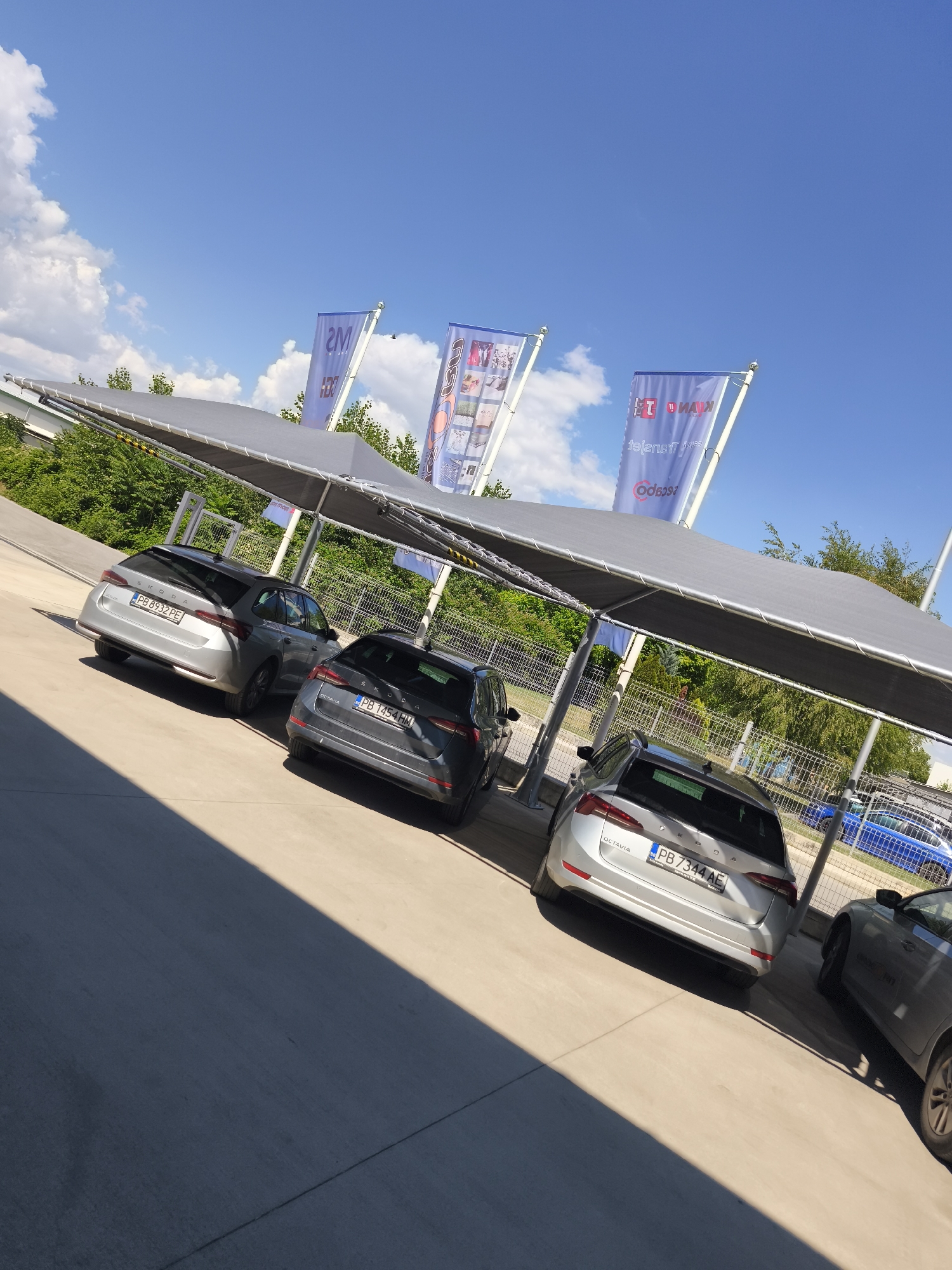 The image shows a car dealership lot on a sunny day. Several cars are parked under a large gray canopy, providing shade. In the foreground, a silver station wagon with license plate PB 8932 PE is visible. To its right, a darker gray station wagon with license plate PB 1454 HM is parked. Further to the right, another silver station wagon with license plate PB 7344 AE is visible. The background shows a chain-link fence, lush green trees, and a bright blue sky with scattered white clouds.
Three large blue flags with white lettering are displayed prominently in the background, adding a corporate or branding element to the scene. One flag on the left appears to have "2M" in large white letters, with smaller text below. The middle flag has a graphic design and some text, while the flag on the right has text that appears to be "MAN TGL" and "MAN TGX" along with a logo.
The lighting suggests it is daytime, likely afternoon, given the strong shadows cast by the canopy and the cars onto the concrete lot. The weather appears to be pleasant and clear, suitable for outdoor activities. The overall scene conveys a sense of commerce and retail, specifically related to automotive sales or services. No people are visible in the frame.