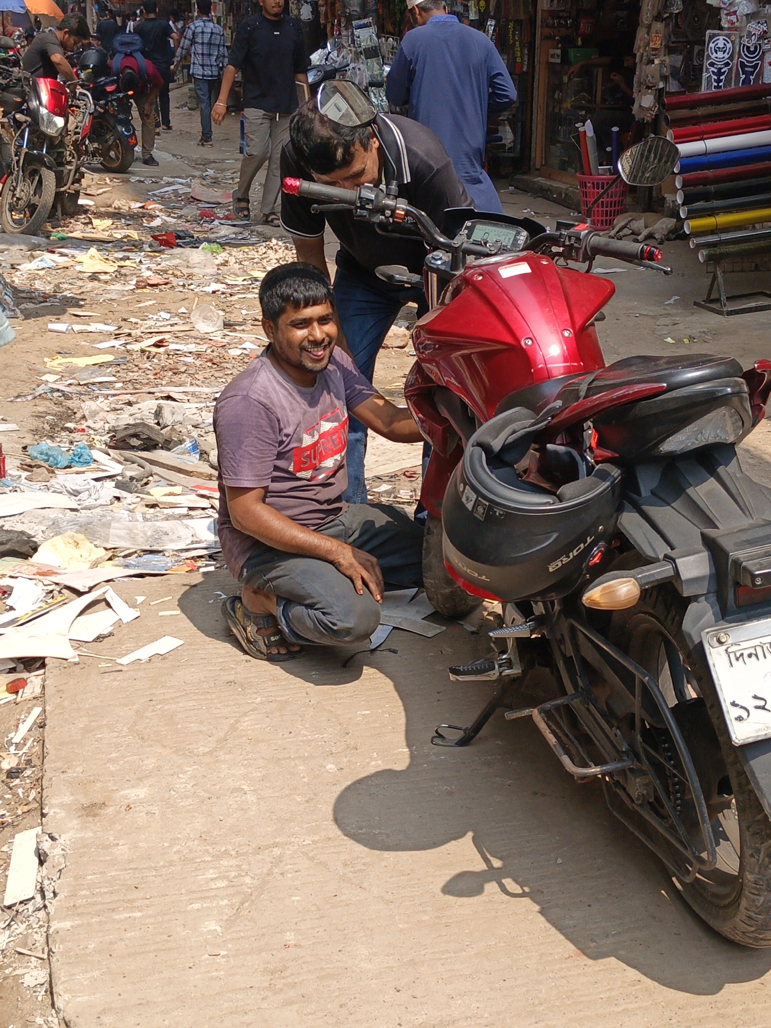 A medium shot captures two men with a motorcycle amidst a busy, cluttered street scene in Dhaka, Bangladesh. The foreground features a man in a purplish-red t-shirt and dark pants, kneeling with a wide smile, his hands visible. Beside him stands a red motorcycle, with a black helmet hanging off the back. The second man, partially visible, is bending over the motorcycle's handlebars.
The street is filled with debris, including scattered papers and trash, suggesting a commercial or market area. In the background, other people and motorcycles are visible, contributing to the bustling atmosphere. A shop is seen lined with rolls of colorful materials, implying a market environment.
