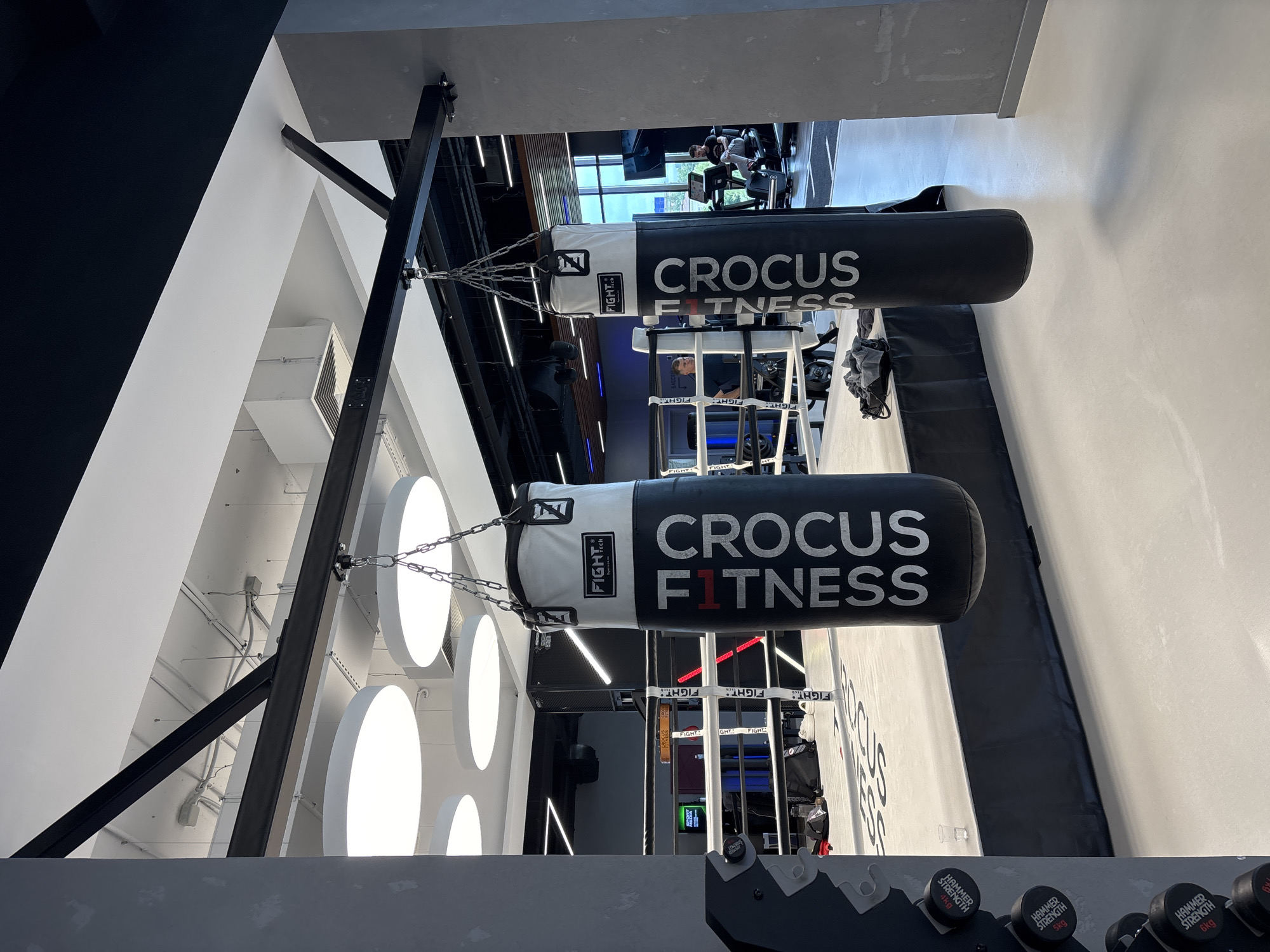 The image depicts the interior of a boxing gym, likely in the city of Monolit, Russia. In the foreground, two large punching bags hang from a metal frame. The bag on the right is predominantly black with white lettering that reads "CROCUS FITNESS." The bag on the left is white with black lettering. A boxing ring is visible in the middle ground, with white ropes and a raised platform. Inside the ring, a person wearing dark clothing is seen standing. In the background, exercise equipment, including treadmills, is present, and several individuals can be observed using the machines. The gym features modern lighting, including circular ceiling lights, and industrial-style exposed beams and ductwork. The overall environment suggests a well-equipped fitness facility.