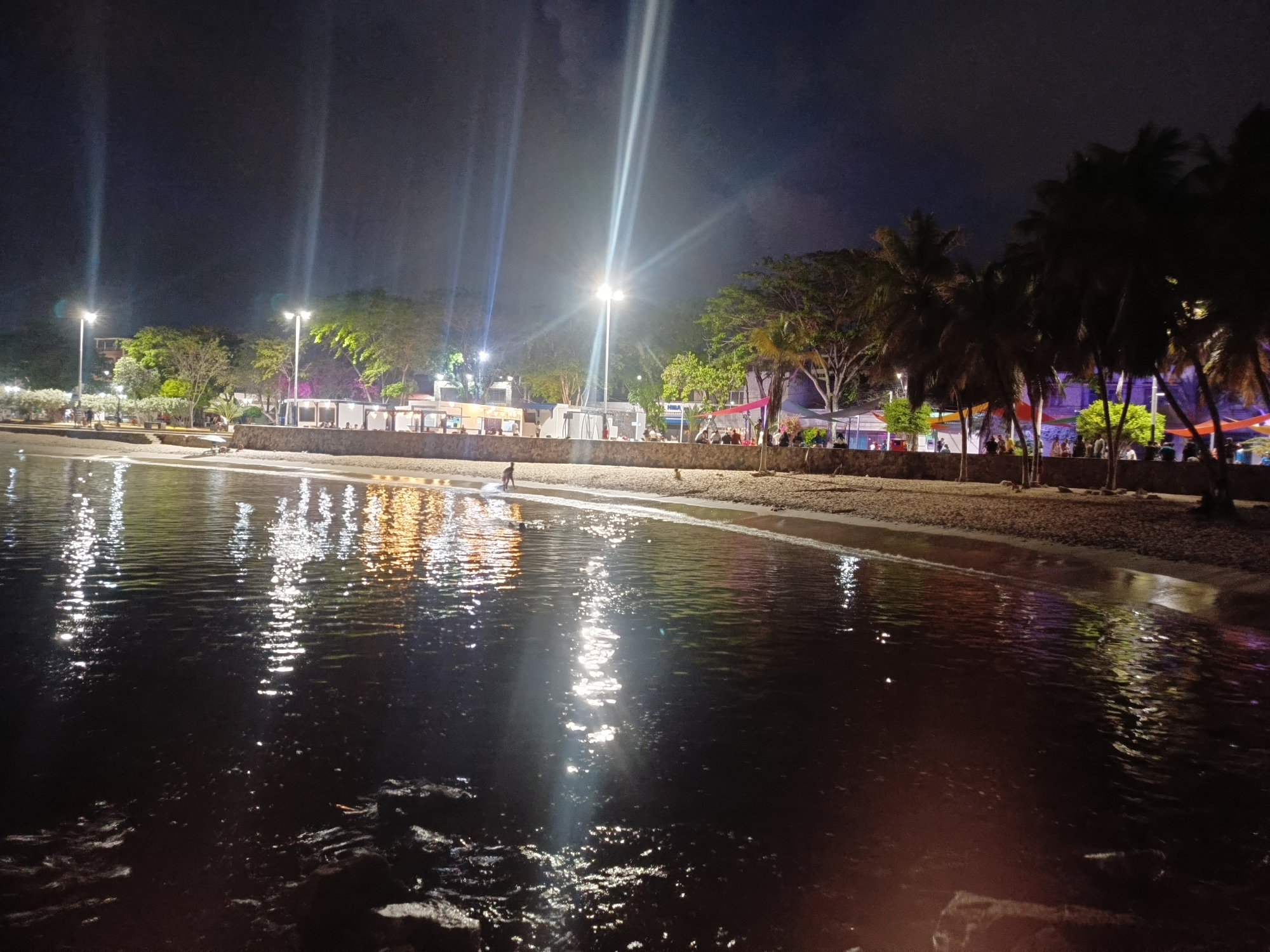 This is a nighttime photograph taken from a beach in Juan Griego, Venezuela, looking towards the shore. The foreground is dominated by dark water, reflecting the lights from the shore. The beach is visible in the mid-ground, with a sand and stone embankment separating it from a promenade or walkway. Several people are present along the shore, some walking and others gathered at what appear to be food stalls or restaurants. Palm trees and other vegetation line the area behind the promenade, and streetlights illuminate the scene. The overall atmosphere is lively, suggesting a popular nighttime gathering spot.