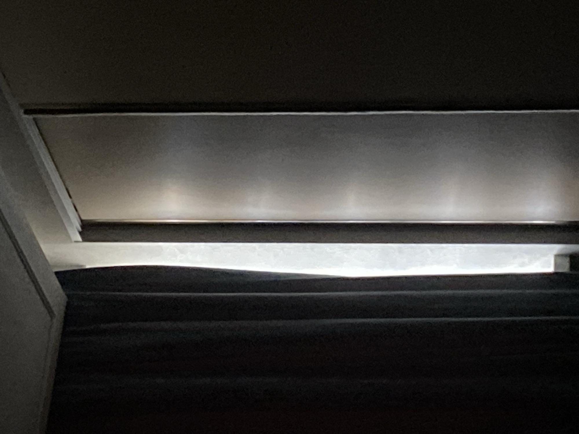 A dark curtain hangs to the left of a narrow, translucent white curtain. To the right of the white curtain is a metallic sliding door with vertical lines. The wall to the right of the door is dark. The scene is lit by light from an unseen source to the left, casting reflections on the sliding door.