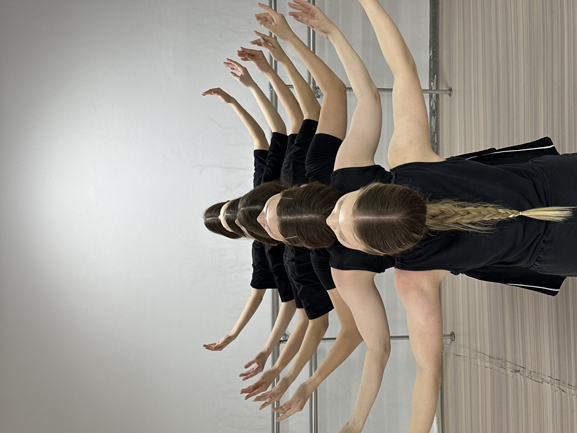 The image shows a row of six images of the same young woman performing a dance move. All are wearing black tops and shorts, with the farthest figure having blonde hair braided with a white tie. The background consists of a white wall on the left and a patterned wood-like surface on the right with a horizontal bar and wall-mounted railing in the middle. The lighting is bright and even. No location is indicated.