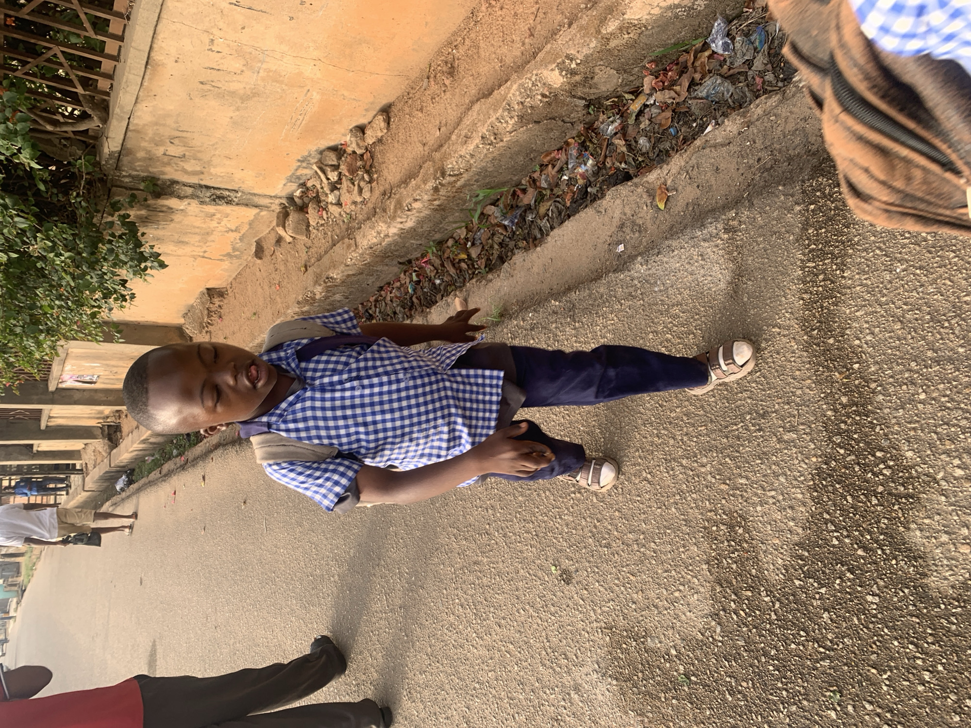 A young child, likely a boy, is the central subject of the image, standing on a light-colored paved road or pathway. He wears a blue and white checkered short-sleeved shirt, which appears to be a school uniform, dark blue trousers, and light-colored sandals. A dark backpack strap is visible over his left shoulder. His eyes are closed, and his mouth is open with his tongue slightly extended, suggesting a playful or expressive moment.
To his right, running parallel to the road, is a concrete gutter or drainage channel filled with dry leaves, soil, and various pieces of litter, including plastic bags and wrappers. Beyond the gutter, there is a low concrete structure, followed by an area with green bushes and some parts of a metal fence or railing. The ground cover consists of sparse vegetation and dirt.
In the background, further down the road, the lower bodies of other individuals are partially visible, one wearing light-colored shorts and another with dark trousers and a red top. The bright sunlight indicates it is daytime, casting subtle shadows. The overall scene depicts an outdoor street environment, possibly in an urban or semi-urban area in Jos, Nigeria, characterized by local architecture and natural elements. There is no visible text in the image.