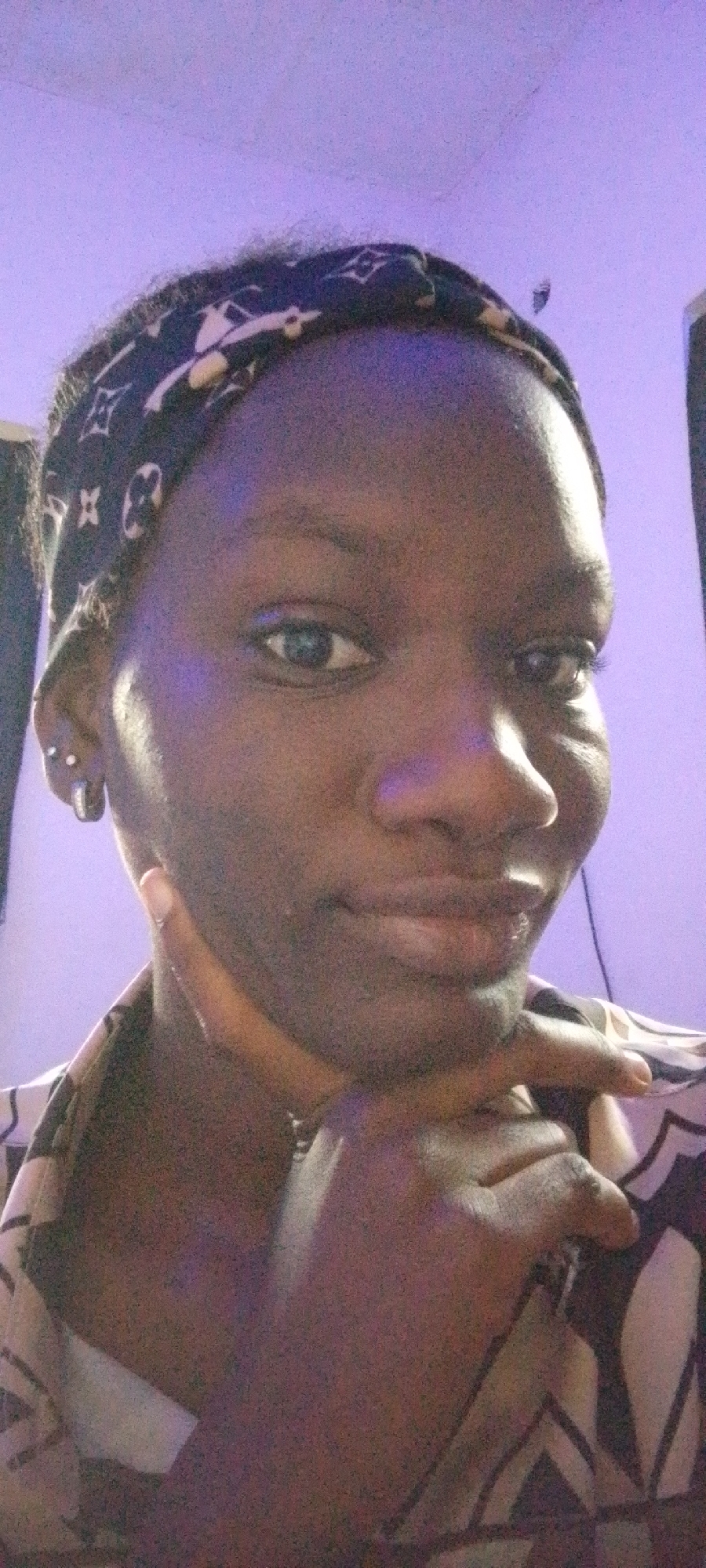 A dark-skinned person is captured in a close-up selfie, their face angled slightly to the right. They wear a black bandana with a repeating pattern of white symbols and a collared shirt with a black and white geometric design. The person's right hand rests on their chin, with their thumb extended towards their cheek. A silver hoop earring is visible on their left ear, along with a smaller silver stud. The background is a softly lit purple wall, suggesting an indoor setting. The lighting is dim, with a slight purplish hue cast over the scene. The overall impression is a casual, introspective moment.