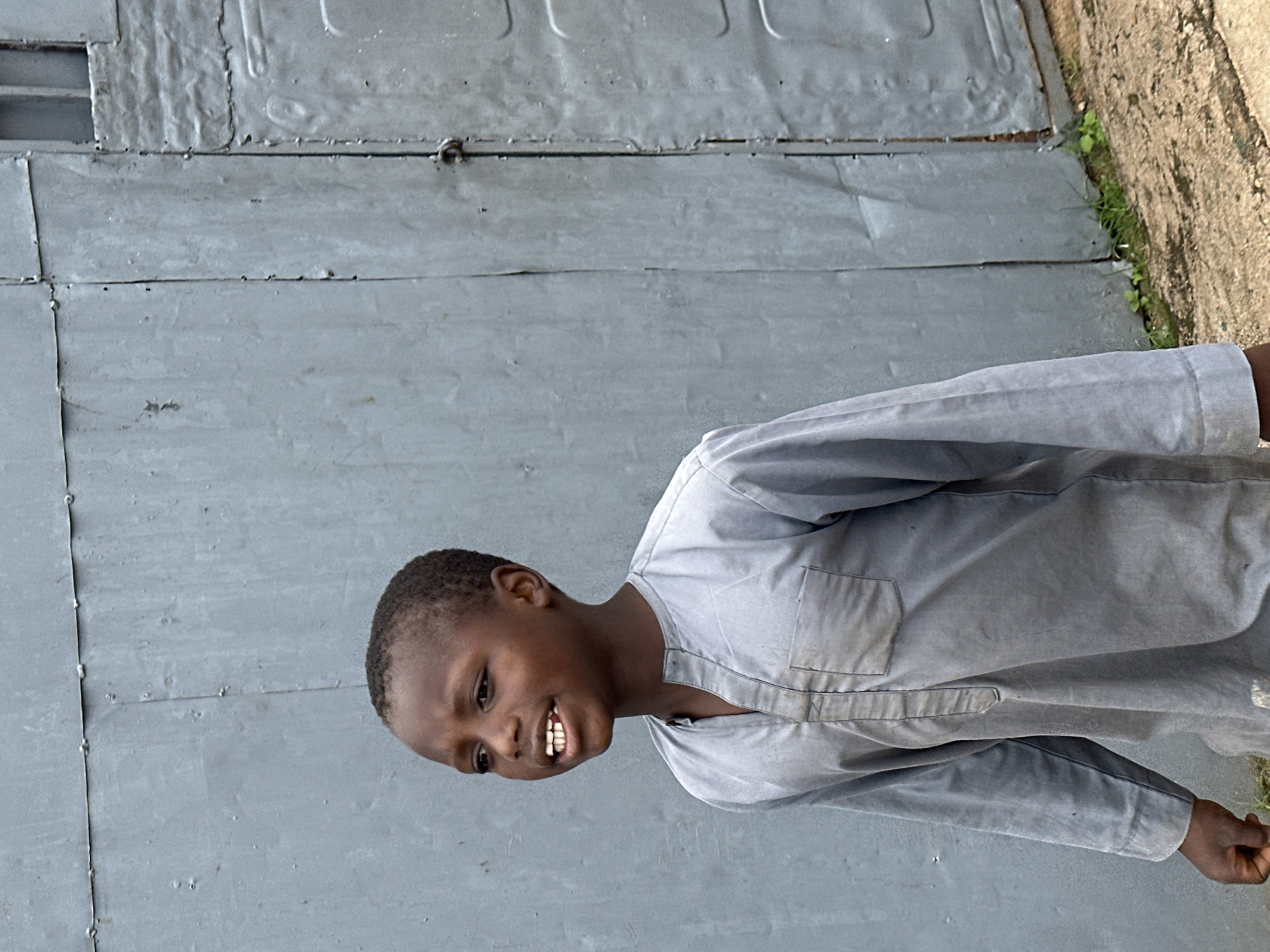 A young boy is pictured in a medium shot, standing in front of a gray corrugated metal wall. He is wearing a gray, long-sleeved tunic with a V-neck and a single pocket on the left side of the chest. His expression is directed to his left, with his mouth slightly open, revealing his teeth. The boy's dark hair is cut short. The metal wall behind him appears weathered and shows signs of rivets and seams. A small patch of green grass and dirt is visible at the bottom of the frame, along the base of the wall. The lighting suggests it is daytime. The scene is devoid of any identifiable landmarks that would definitively place it in Jos, Nigeria, beyond the user's input.