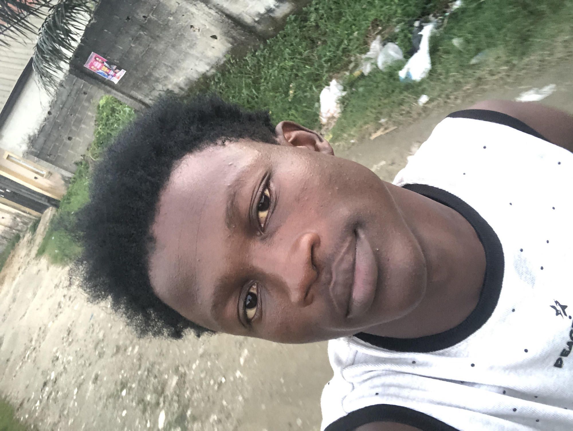 The image is a selfie taken by a young Black male with dark, curly hair, wearing a white sleeveless shirt with a black trim. He is outdoors with a dirt and grass path behind him. To his right, there is a gray concrete wall with some graffiti or posters and green vegetation. A portion of a building with yellow trim is visible on the far left. The lighting suggests it is daytime. The background is somewhat out of focus, with a few pieces of white trash visible on the ground. The setting appears to be an urban or semi-urban environment, consistent with a location in Ado, Nigeria.