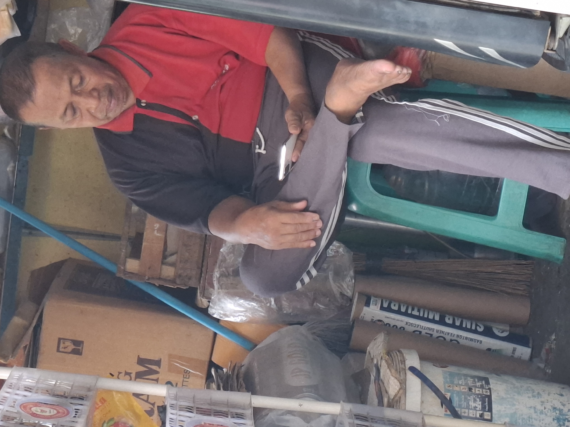 The image shows a man sitting on a green plastic stool in what appears to be a storage area or small shop. The man is wearing a two-toned polo shirt, with one side black and the other red, and grey track pants with white stripes down the sides. He is holding a smartphone in his right hand and looking down at it. His left leg is bare and rests on the stool.
Behind the man, there are stacks of boxes, including one with a fragile symbol. There are also several large cardboard tubes, some wrapped in plastic, and a broom leaning against them. To the left of the man, there are white plastic shelves filled with various items, including what looks like packaging and possibly food products. A blue pipe runs vertically in the background. The overall environment suggests a cluttered storage space. The lighting is somewhat dim, with a yellow-toned light source. The setting is consistent with a shop or storage area in a densely populated urban environment like West Jakarta, Indonesia, characterized by the presence of various goods and storage solutions.