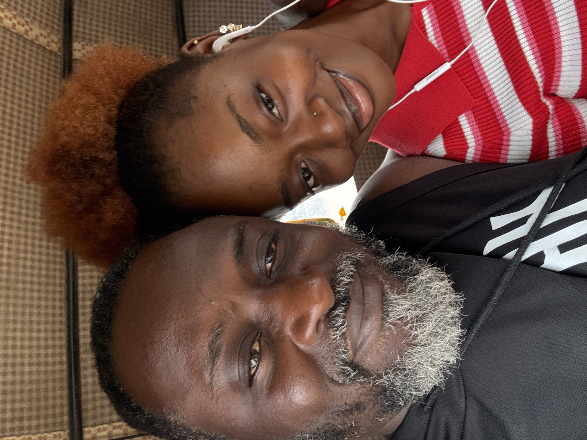 Two individuals are captured in a close-up, selfie-style photograph, with their faces angled towards the camera. The background appears to be the interior of a vehicle, possibly a bus or keke, indicated by the checkered fabric upholstery and a hint of a window or roof lining. The lighting suggests an indoor environment with natural light sources.
On the left is a man with dark skin, a greying beard and mustache, and dark, short hair. He is wearing a black hooded jacket with white lettering on the chest. His gaze is directed at the camera, and he has a slight smile.
On the right is a young woman with dark skin and reddish-brown hair styled in an afro. She is wearing a red and white striped collared shirt. She has on white earbuds and subtle jewelry on her ear and nose. She is smiling broadly and looking towards the camera. Her left arm is around the man's shoulder, indicating an affectionate or familiar relationship. The interaction between the two suggests a shared moment of closeness and enjoyment.
While the image itself does not explicitly denote the location as Ado, Nigeria, the context of the interior of a public transport vehicle and the general appearance of the individuals are consistent with urban settings in Nigeria.