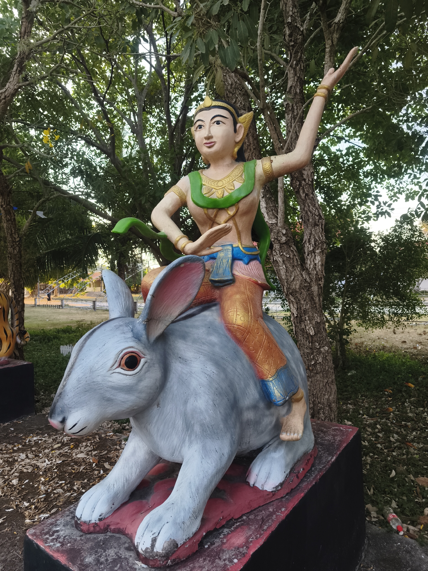A vibrant statue of a human-like figure riding a large rabbit is the central focus, set outdoors in Baan Bo Phut, Thailand. The figure, depicted with golden skin, wears an elaborate golden headdress, a green and gold top, and orange pants accented with blue. It has distinct pointed ears and is captured in a dynamic pose with one arm gracefully raised and the other bent at the waist, indicative of a dance or traditional gesture.
The figure is seated upon a sizable grey rabbit statue, which features reddish-pink eyes and pink inner ears. The rabbit rests on a dark base with red highlights. The scene is enveloped by lush greenery, with numerous trees forming a dense canopy that filters the daylight, suggesting it is daytime. The ground beneath the statue is covered in dirt and scattered dry leaves. In the background, beyond the immediate foliage, there are glimpses of an open grassy area and distant structures, possibly white fences or scaffolding, hinting at a larger public park or temple complex. There are no activities taking place, as the image captures a static monument. No visible text is present.