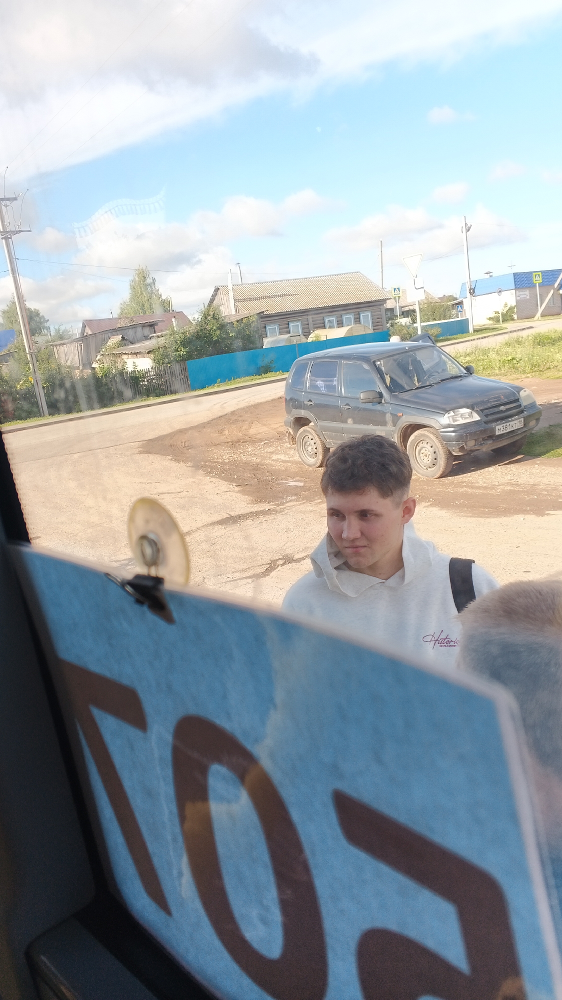 A young man with short, dark hair, wearing a light gray hooded sweatshirt, is looking to his right with a neutral expression. He is standing in front of a dark-colored SUV parked on a dirt and gravel area. Behind the SUV, there are several wooden houses with pitched roofs, some with visible windows, and a bright blue fence. The environment appears to be a rural or village setting. A sign with large, dark lettering on a light blue background is partially visible in the foreground, obscuring the lower portion of the image. The sky is blue with scattered white clouds. The license plate on the SUV is partially visible and appears to be in a Cyrillic script. The location is identified as Kiyasovo, Russia.