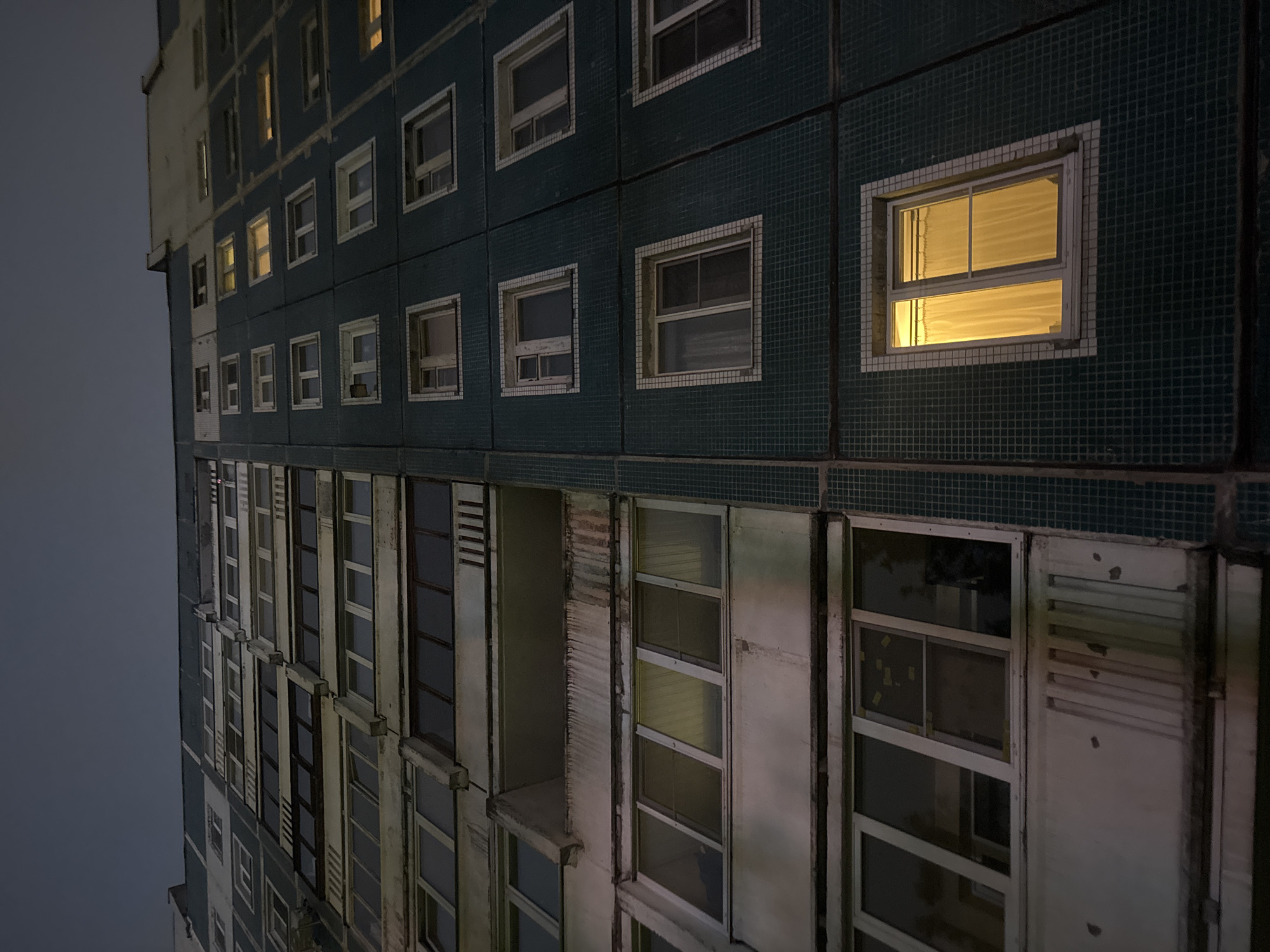 A low-angle shot captures a building in Unknown, Unknown at dusk or night. The building is characterized by its varying exterior styles, with the top portion having a dark teal tiled façade punctuated by square windows, some of which emit a warm, yellow light. The lower part of the building has a lighter, weathered appearance, marked by rows of vertically oriented windows and areas of visible deterioration. The sky above is an overcast gray, providing minimal light.