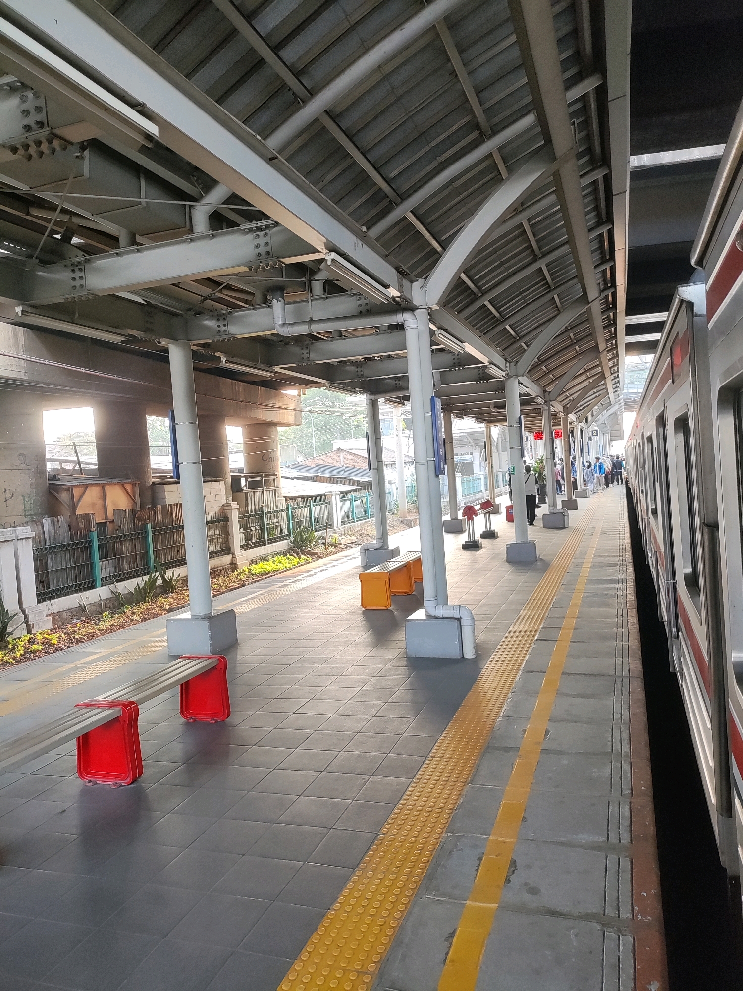 The image shows a train station platform in Bekasi, Indonesia. A modern train with red and white stripes is stopped at the platform. The platform itself is paved with gray tiles and features a yellow tactile warning strip along the edge. There are several modern benches with red legs on the platform. Overhead, a metal canopy supported by white pillars provides shelter. In the background, a concrete overpass is visible, with some low-rise buildings and greenery beyond. Several people are standing on the platform, waiting for or disembarking from the train. The scene appears to be captured during daylight hours, with sunlight casting shadows on the platform.