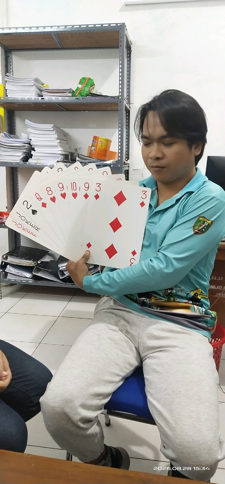 A man is sitting on a chair, holding a fan of oversized playing cards. He is wearing a light blue long-sleeved shirt and light grey sweatpants. The playing cards are large and have red diamond symbols on them. Behind the man is a metal shelf unit filled with books and other items. The floor is tiled. The image appears to have been taken indoors.