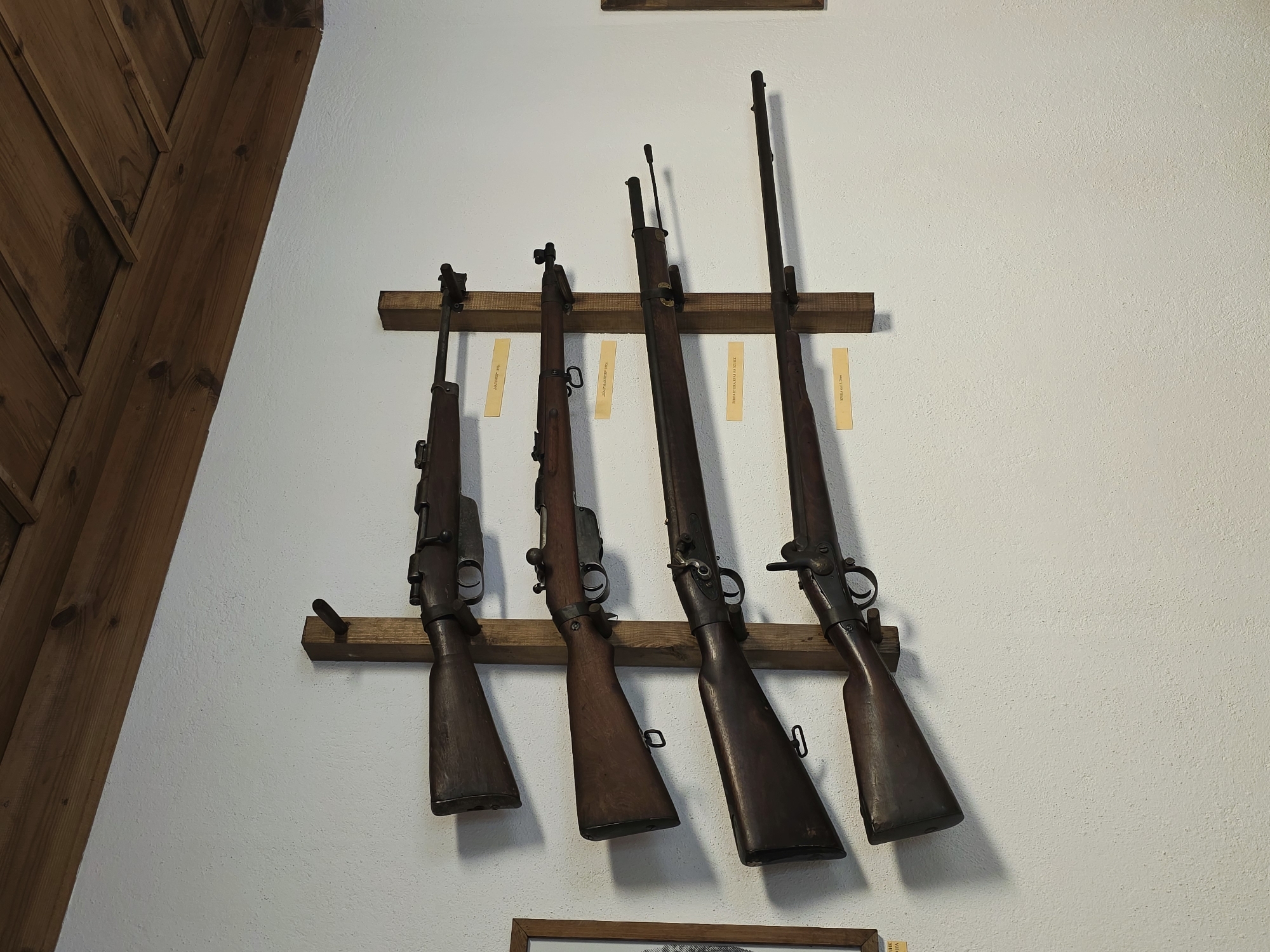 The image shows four rifles mounted on wooden racks against a white textured wall. The rifles are positioned vertically, one above the other, with the butts of the rifles facing downwards and the barrels extending upwards. Each rifle is made of wood and metal, with varying designs and lengths. Small yellow labels are attached to the wall beneath each rifle, presumably indicating their origin or type. Above the rifles, a wooden ceiling structure made of planks is visible. The overall scene suggests a display of historical firearms, possibly in a museum or collection. The provided crop images offer closer views of the rifles, their wooden stocks, metal mechanisms, and the labels. There are no people visible in the image. The environment appears to be indoors, with artificial lighting and a rustic aesthetic. Kovachevitsa, Bulgaria is a village known for its preserved architectural heritage, and such a display of historical artifacts would be fitting within its context.