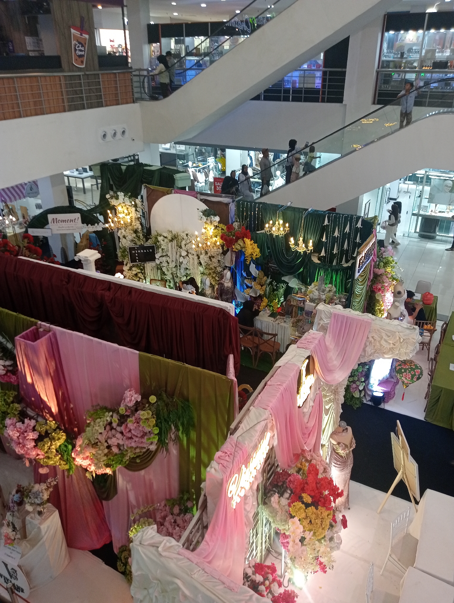 The image shows an indoor scene, likely a shopping mall, with multiple levels and escalators. In the foreground, there is a decorative display featuring abundant floral arrangements in shades of pink, red, yellow, and white, draped fabrics in pink and green, and illuminated signage. A mannequin in a formal gown stands near the floral display. In the middle ground, more elaborate decorations with green drapery, chandeliers, and floral arrangements are visible. Further back, people are seen on escalators and walking on the upper levels. The environment appears to be a commercial setting with shops and displays. The presence of the "Chatime" logo on the upper level suggests a commercial establishment. The setting is indoors with artificial lighting. The style of the decorations suggests a festive or celebratory event, possibly a wedding or similar occasion. The mention of Jambi, Indonesia, implies this scene is located in that city.