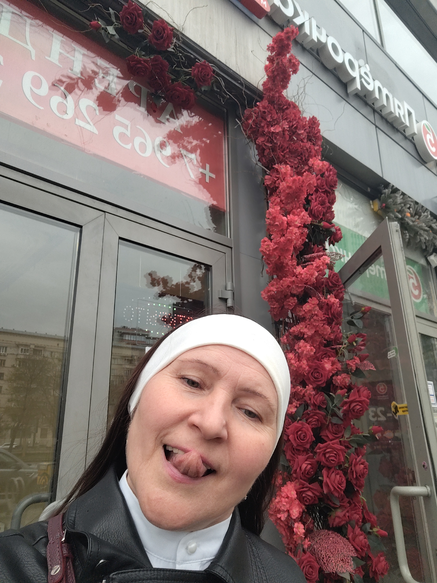 A middle-aged woman with dark hair, wearing a white headband, a white collared shirt, and a black leather jacket, is taking a selfie in front of what appears to be a storefront decorated with red flowers. She is sticking her tongue out playfully. The building has signage in Cyrillic. The storefront's sign reads "Пятёрочка" (Pyaterochka), which is a Russian chain of grocery stores. The location is in Moscow, Russia.