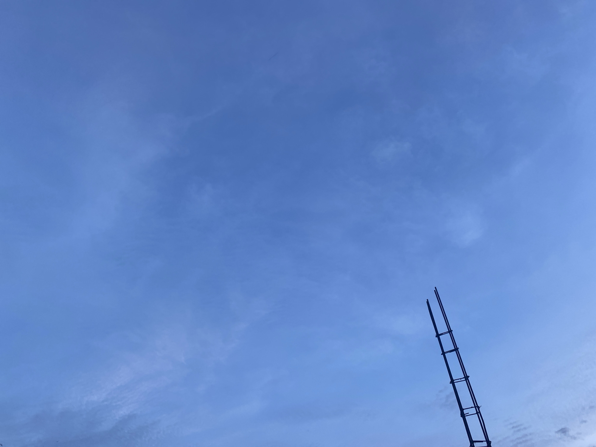 The image depicts a clear blue sky with wispy clouds, and a dark silhouette of a ladder extending upwards from the bottom right corner. The ladder is positioned diagonally, with its top reaching towards the upper right of the frame. The sky is predominantly a deep blue, with lighter shades closer to the horizon, suggesting either early morning or late evening. There are no people visible in the image, nor any other objects or structures that would provide context for the ladder's presence or location. The scene appears to be an outdoor environment, likely in Banda Aceh, Indonesia, given the prompt's specification. The overall impression is one of an open, unobstructed view of the sky, with the ladder serving as the only prominent man-made element.