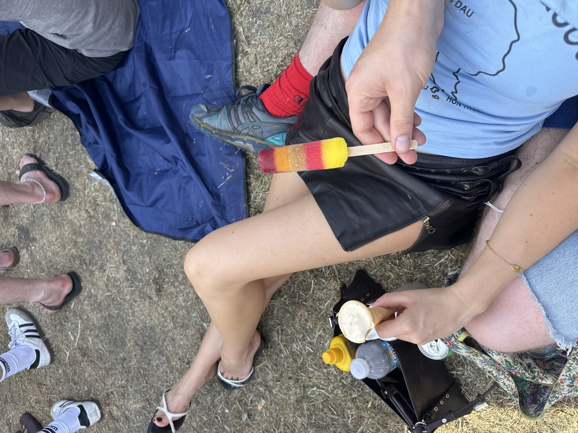 The image shows a group of people gathered on a grassy surface, possibly a park or field. One person is holding a colorful popsicle, while another is holding an ice cream cone with a bite taken out of it. There is a water bottle, a can, and a yellow container (possibly mustard) near the ice cream cone. Some of the people are sitting on a blue blanket. Various footwear is visible, including sandals, sneakers, and athletic shoes with red socks. A purse or bag is nearby. The setting appears to be outdoors on a sunny day. There is no clear evidence to confirm the location is London, United Kingdom.