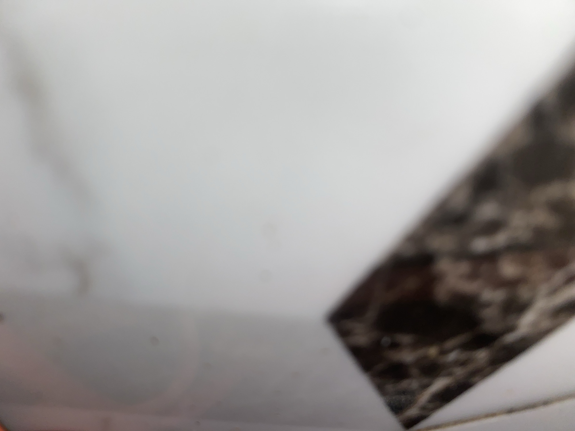 The image shows a close-up, out-of-focus view of a white surface with a dark, patterned diagonal band. The patterned band appears to have a marbled or textured appearance with shades of brown and black. The white surface takes up the majority of the frame and is smooth and featureless, with some slight variations in lighting and subtle darker smudges. The overall impression is that of a flat, horizontal surface, possibly a countertop or floor. There are no discernible people, objects, or actions taking place. The location in Jauro Yinu, Nigeria, cannot be determined from the visual information provided.