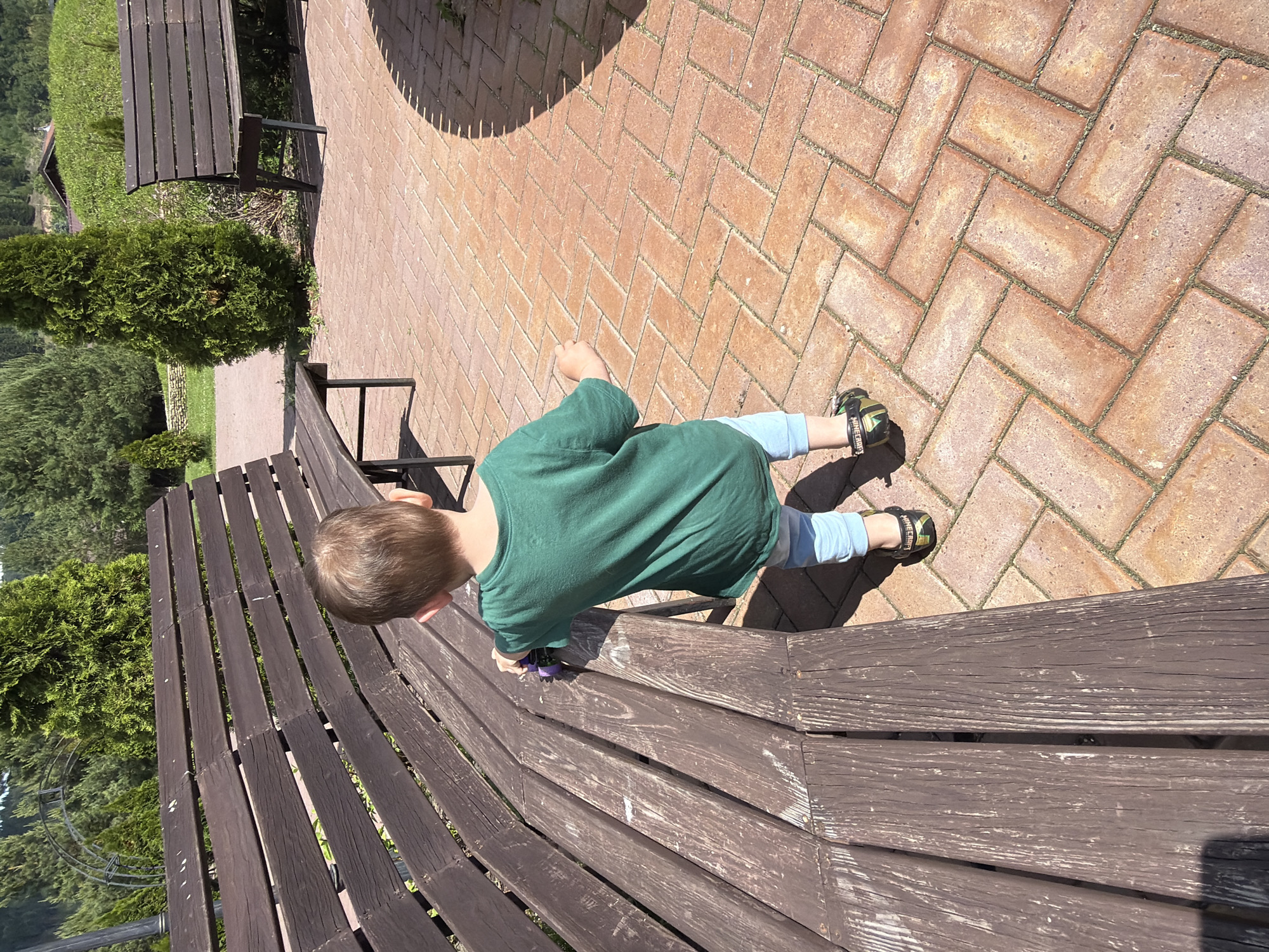 A young child, facing away from the camera, stands on a herringbone-patterned brick patio next to a weathered, dark brown wooden park bench. The child is wearing a green t-shirt, light blue pants, and black sandals with a green accent and the word "MINECRAFT" visible on the side. The child's left hand is touching the bench. In the background, there are more wooden benches, green trees, and a glimpse of a building and hillside. The scene appears to be outdoors in a park or public space. The location is identified as Красный Ключ, Russia.