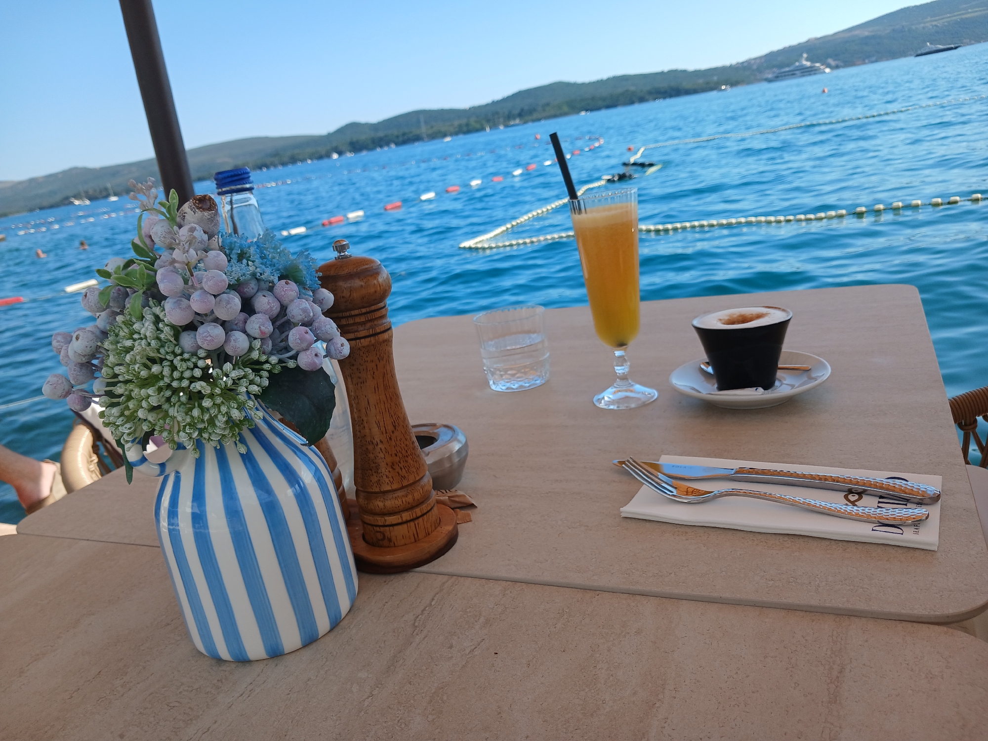 On a table overlooking a body of water, a vase with a floral arrangement, a salt and pepper grinder, a glass of water, a glass of orange juice with a straw, and a cup of coffee are arranged. Cutlery is set on a napkin to the right of the table. The body of water has buoys and a few boats in it, and hills can be seen in the distance under a clear blue sky.