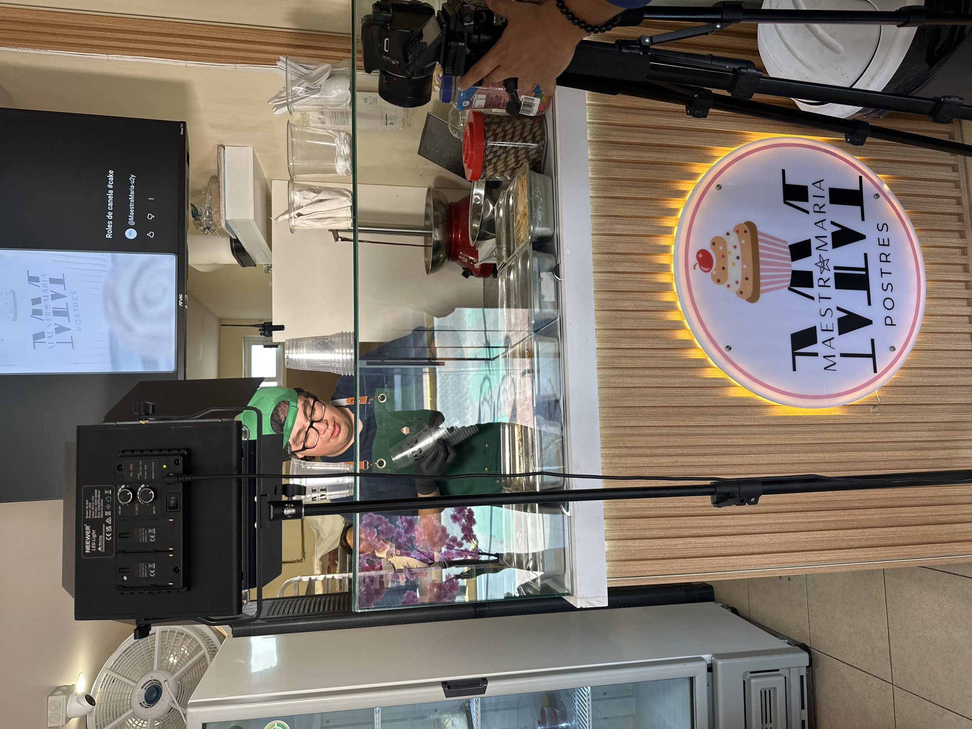 Here is a detailed description of the image:
**Scene:** This indoor shot appears to be taken at a pastry business called "TATAL MAESTRA MARIA POSTRES," located in San Andrés Cholula, Mexico. The image captures what seems to be a filming setup in front of a pastry display case.
**People:** A person wearing a green cap, glasses, and an apron is visible behind the display case, possibly preparing pastries. Another person is partially visible, holding a professional camera (DSLR or similar).
**Subjects:** The primary subject seems to be the preparation or display of pastries. A television screen in the background shows the business' logo, and the text "Roles de canela #cake," and "@MaestraMaria-u2y"
**Objects:**
* Pastry display case with various utensils and containers.
* A NEEWER LED light with batteries attached.
* A fan situated below the LED light.
* A sign with the business name and a cupcake image.
* A tripod.
* A refrigerator.
**Actions:** The person behind the display case appears to be working on something, possibly preparing pastries. The person holding the camera seems to be filming.
**Interactions:** The interactions mainly involve setting up a video shoot to showcase the pastry business, potentially for promotional content.
**Environment:** The location is a well-lit pastry shop. The walls are off-white with wood panelling. The lighting suggests a professional setup to enhance video or photo quality.