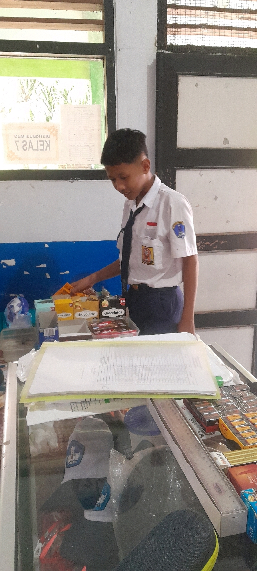 A young male student, wearing a white collared shirt, dark tie, and navy blue pants, stands at a counter. He is reaching for a yellow box of "Chocolatos" snack bars, which is stacked with other similar snack boxes and some chocolate bars in a clear plastic container. In front of the snacks, there are papers and folders on the counter. The background shows a white wall with a blue stripe, and windows with a view of vegetation outside. The scene appears to be indoors, possibly a classroom or a small shop, given the presence of school-related items like the emblem on the student's shirt and a ruler visible on the counter. The overall impression is one of a casual, everyday moment.